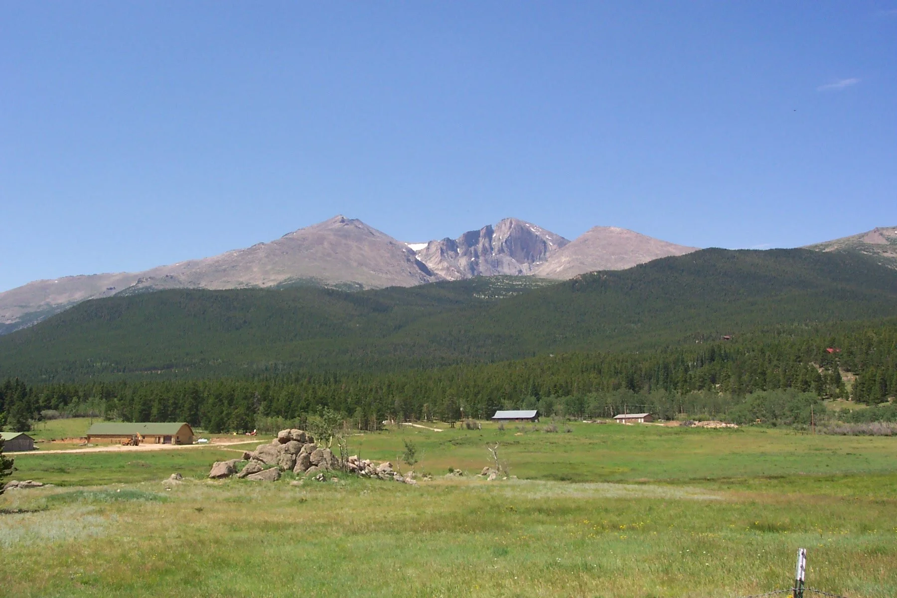 A lush green field with scattered rocks and small farm buildings in the foreground, a dense forested hillside in the middle ground, and towering rocky mountains under a clear blue sky in the background.