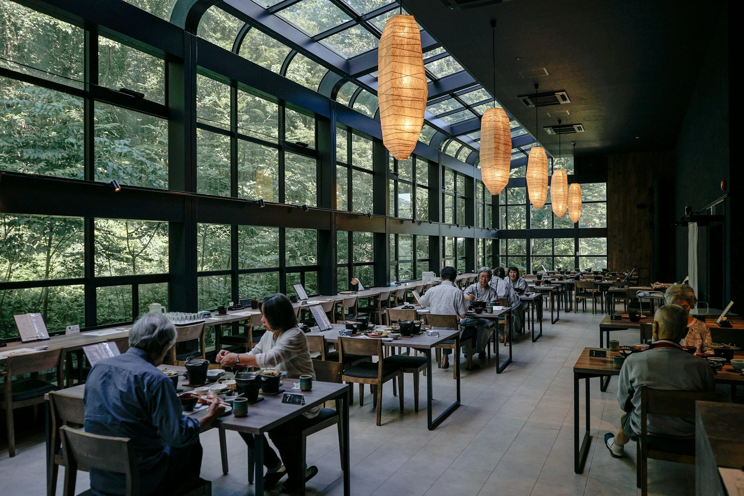 Interior of a modern restaurant with large glass windows overlooking greenery, hanging paper lantern lights, and patrons dining at tables.