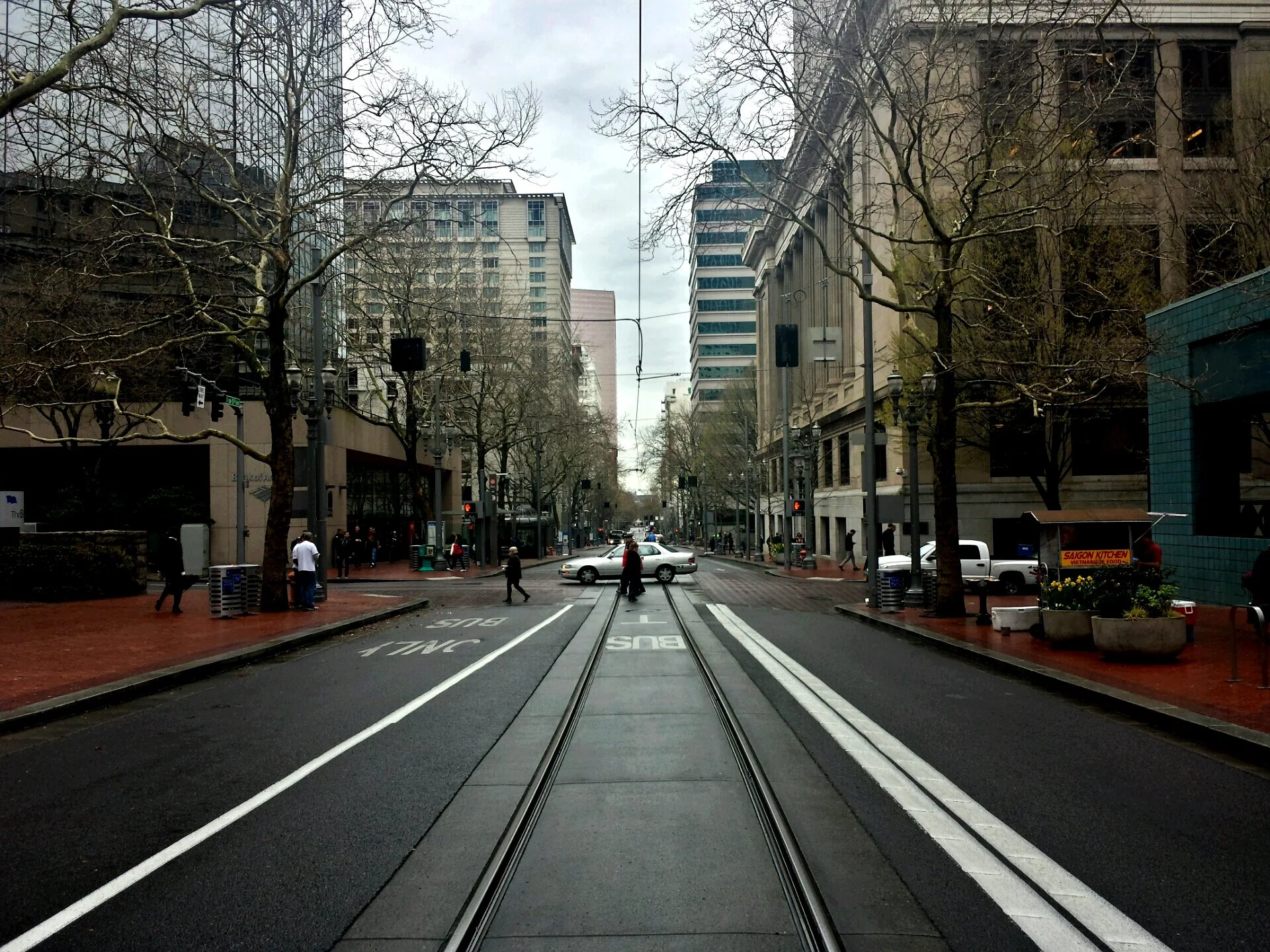 City street with buildings on both sides, leafless trees, pedestrians, cars, and tram tracks in the middle of the road.
