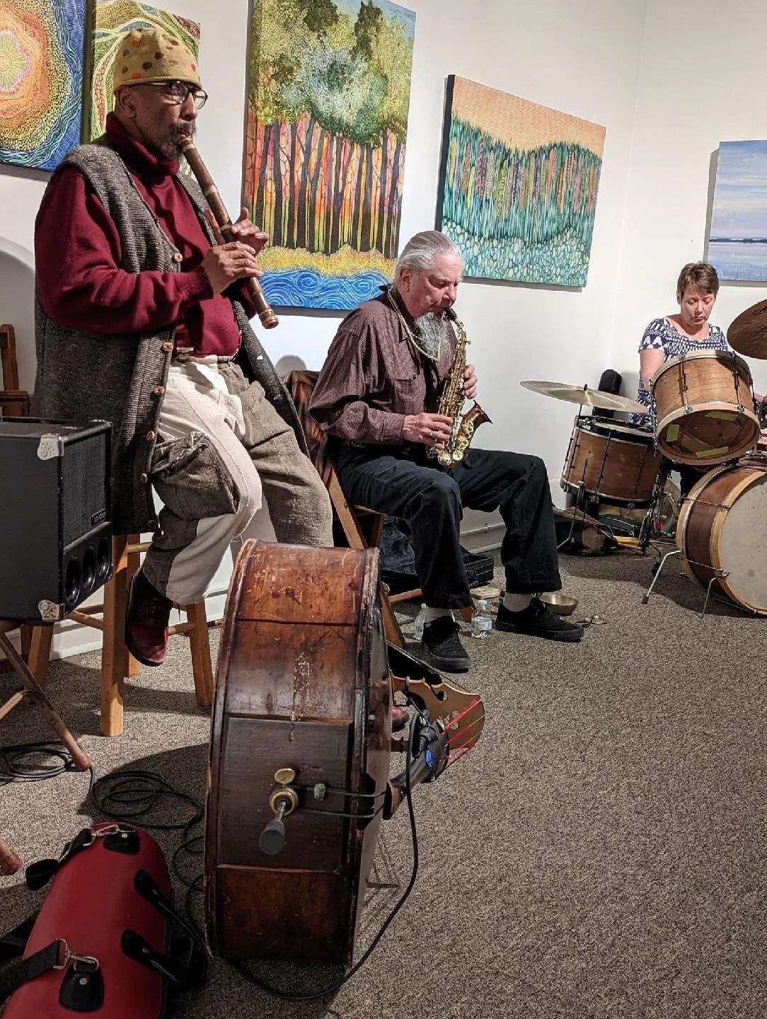 Three musicians performing in an art gallery, playing a clarinet, saxophone, and drums, with colorful paintings on the wall behind them.