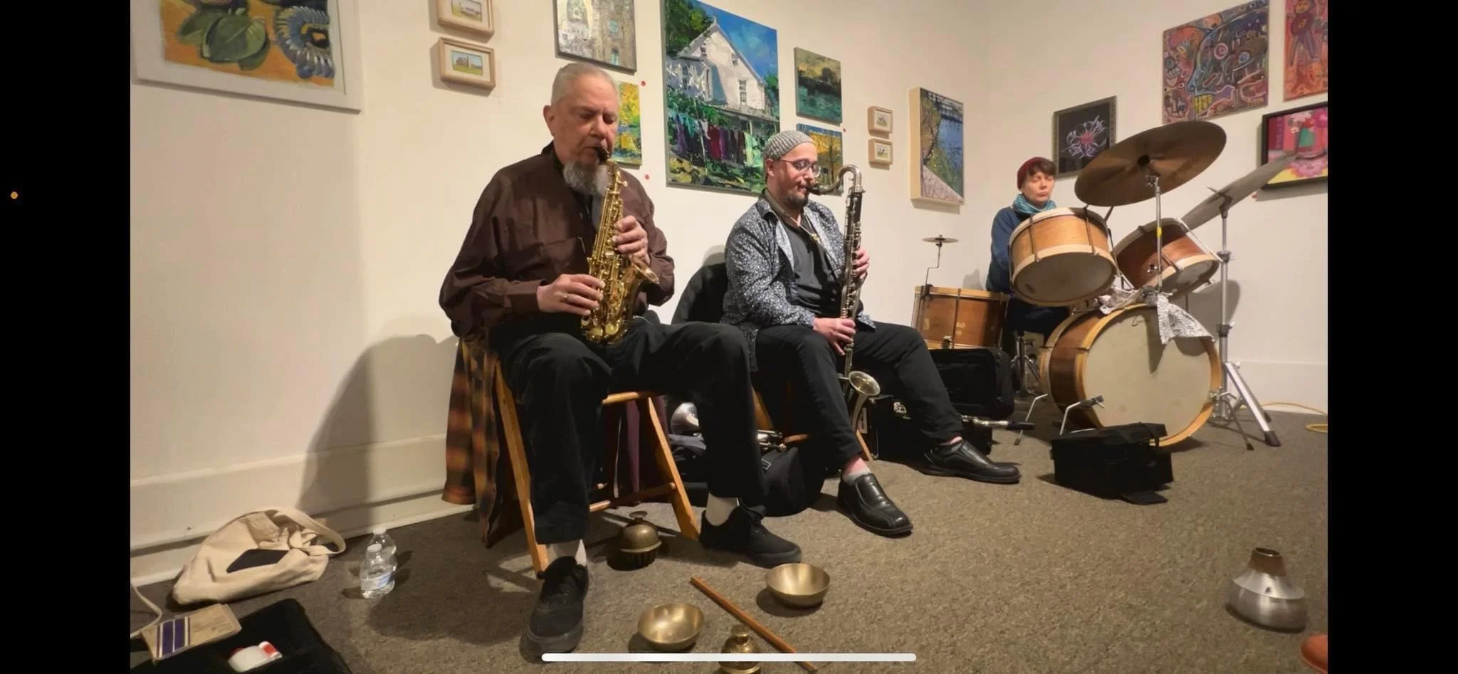 Three musicians performing indoors with a wall of colorful paintings behind them. The first is playing a saxophone, the second a clarinet, and the third is at a drum set. There are musical instruments and personal belongings on the floor in front of 