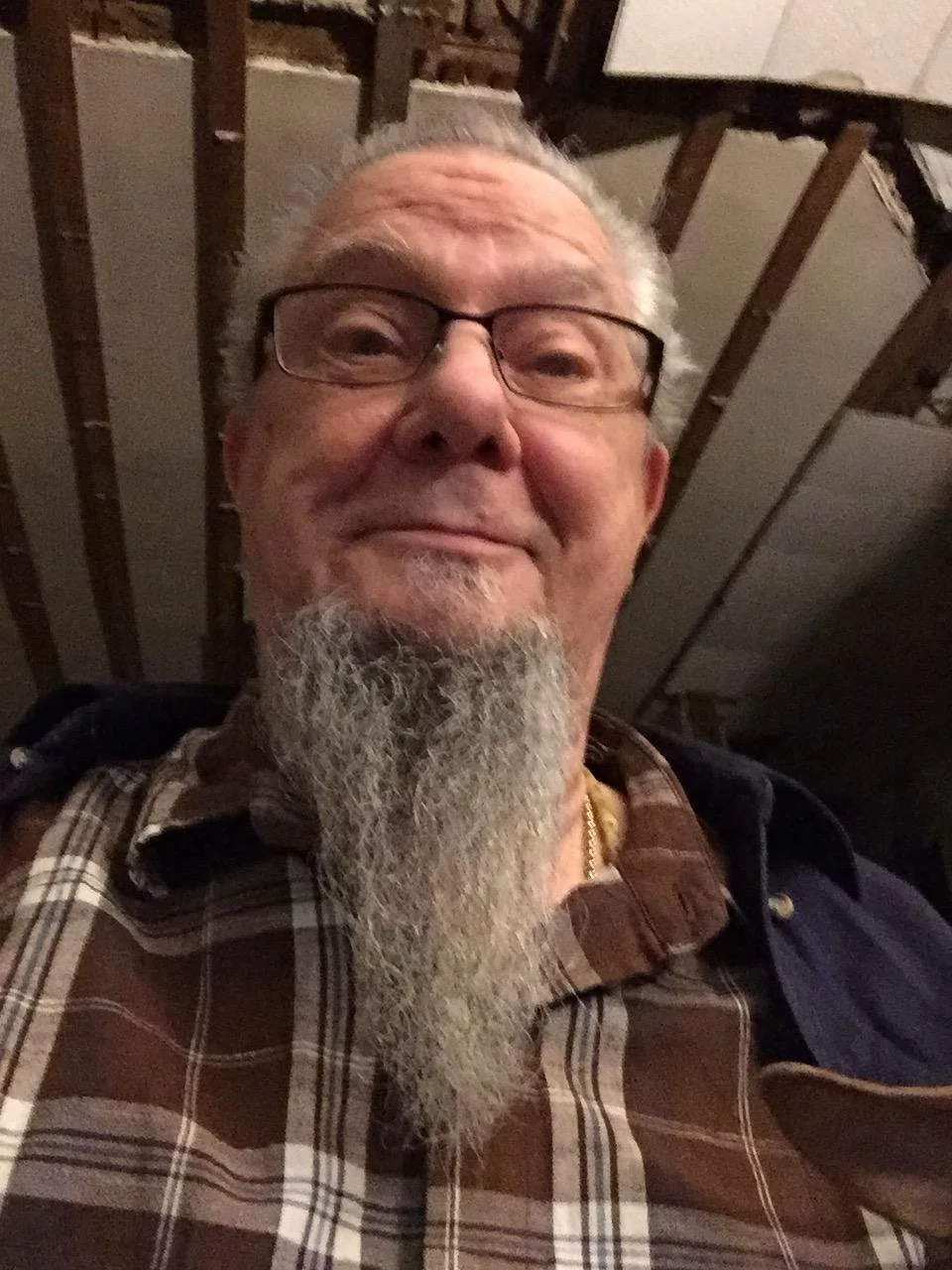 A close-up of an older man with glasses and a long gray beard, smiling at the camera, in an indoor setting with exposed ceiling beams.