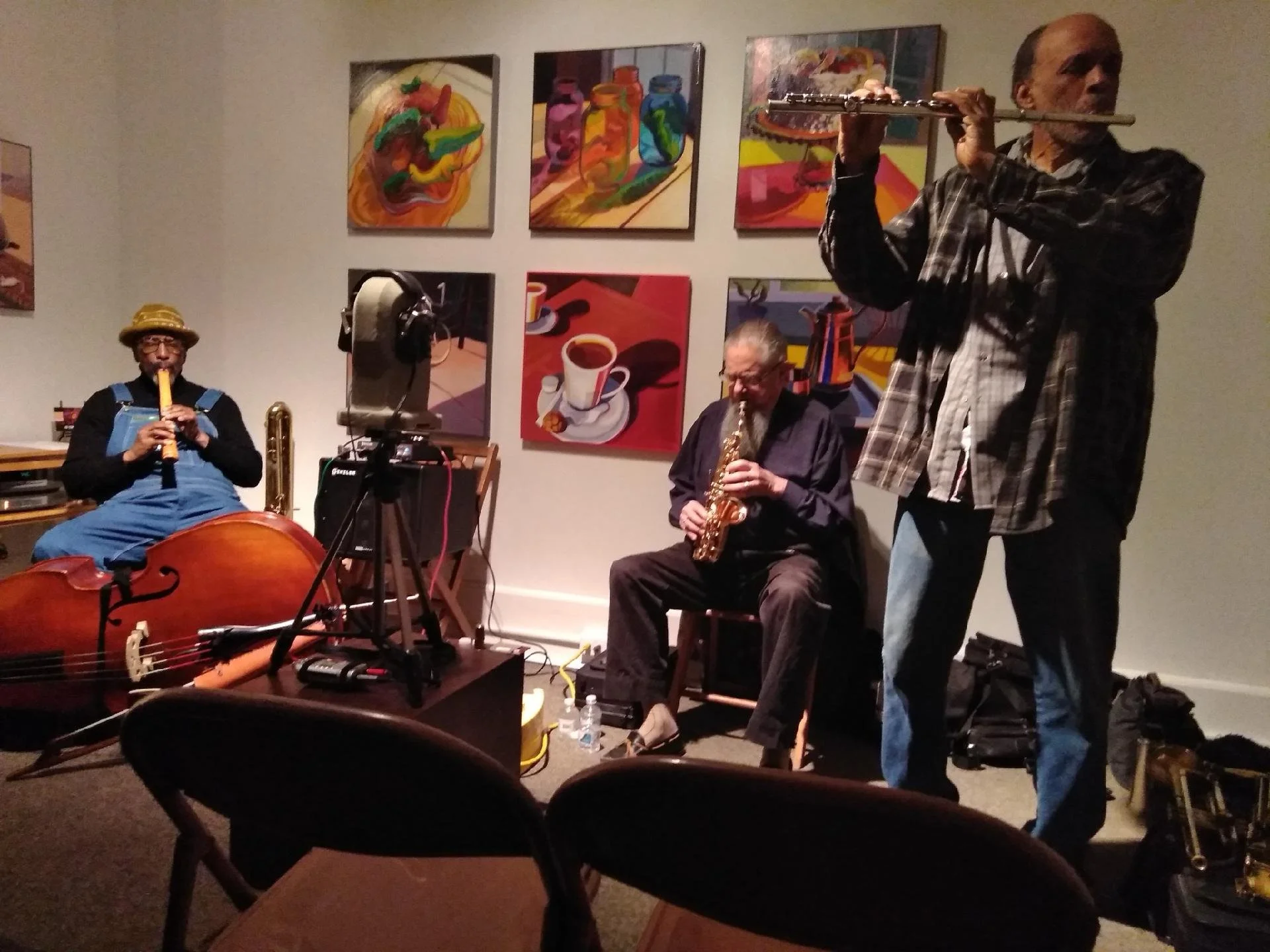 Three musicians performing in an art gallery. One playing a recorder, another on a saxophone, and the third on a flute. Art paintings decorating the wall behind them.