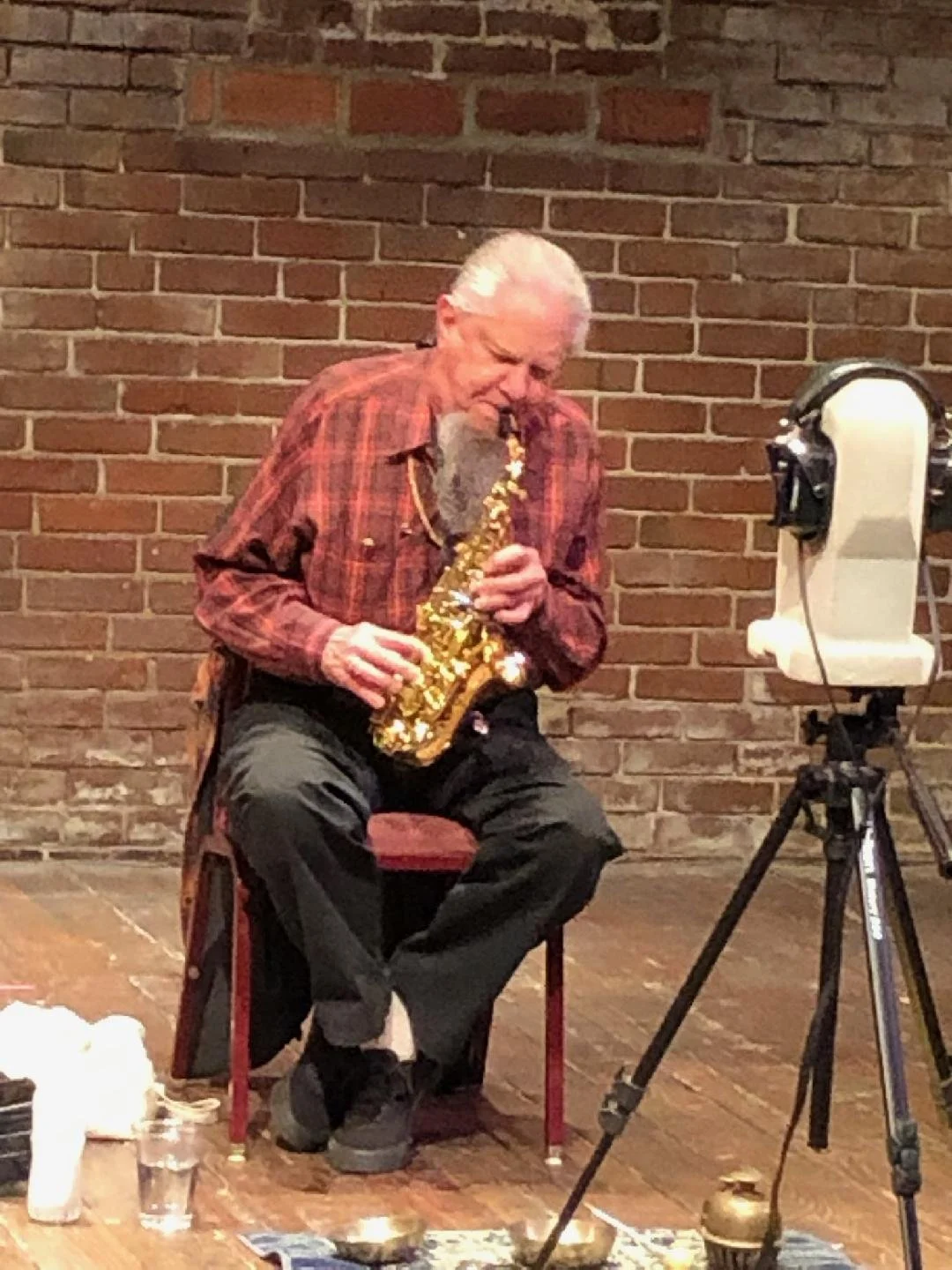 An elderly man with white hair, wearing a red plaid shirt and black pants, seated on a red stool, enthusiastically playing a gold-colored saxophone in front of a brick wall, with a camera on a tripod recording him.
