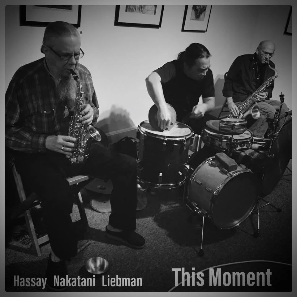 Three musicians playing jazz instruments: a saxophonist, a percussionist, and a saxophonist, in a room with framed artwork on the wall. The photo is in black and white, and the text reads "Hassay Nakatani Liebman" and "This Moment."