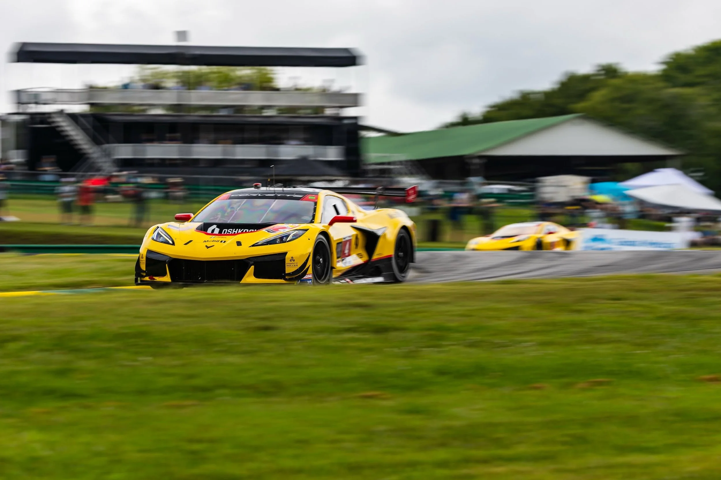 Yellow Corvette race car cornering at speed on a racetrack, with spectators and VIR track facilities in the background.