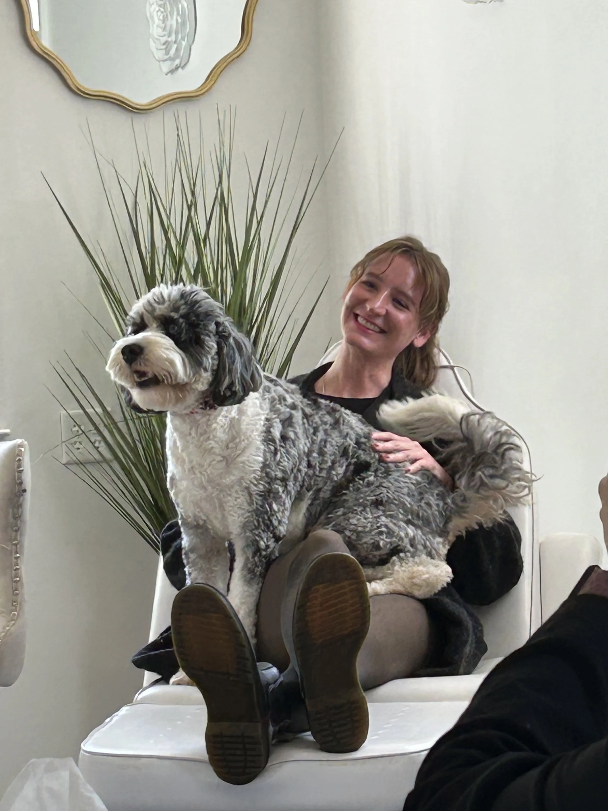 A man performing chiropractic adjustment on a woman seated in a chair, with a dog standing nearby in a room with framed artwork on the wall.
