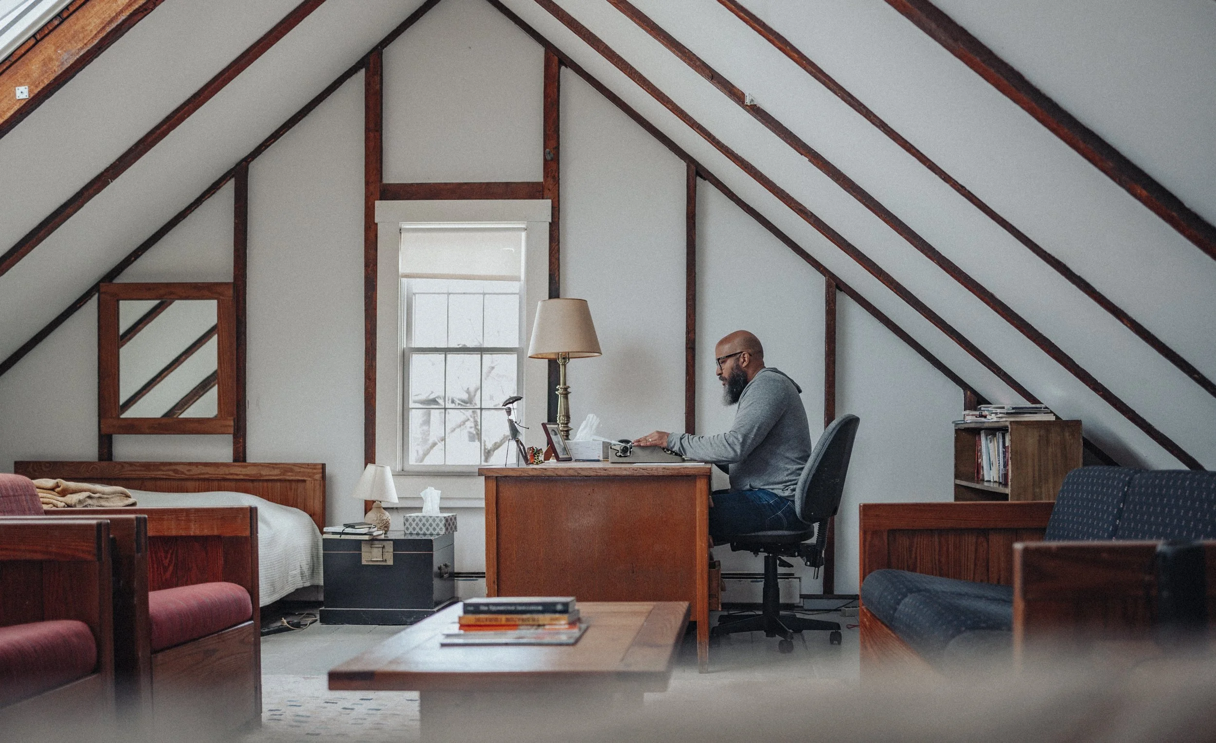 Man working at a desk in an attic office with sloped ceilings, featuring wooden furniture, a bed, and a window.