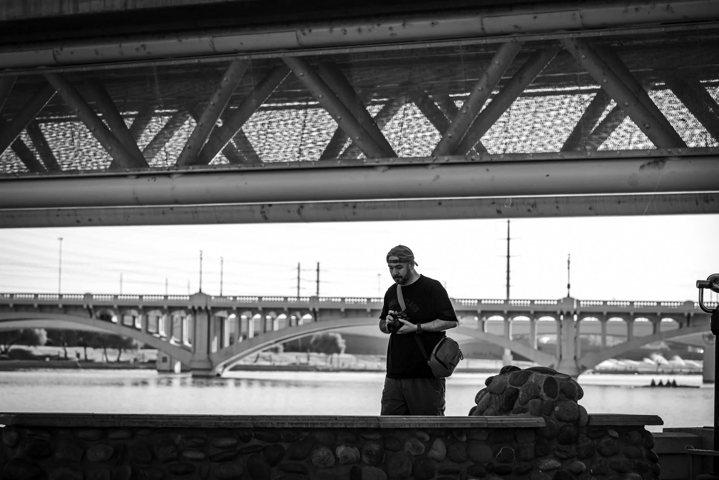 A man in a baseball cap standing on a bridge near a river, holding a camera, with multiple bridges and power lines visible in the background.