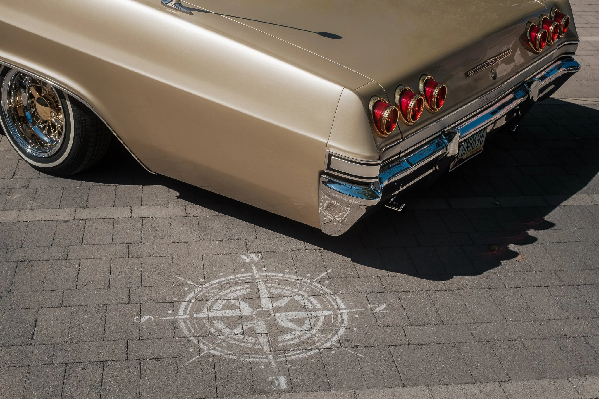 The rear view of a vintage beige and gray car with chrome details, parked on a brick pavement. The car features multiple round tail lights and a European license plate. There is a white circular symbol painted on the ground below the car's rear bumper.