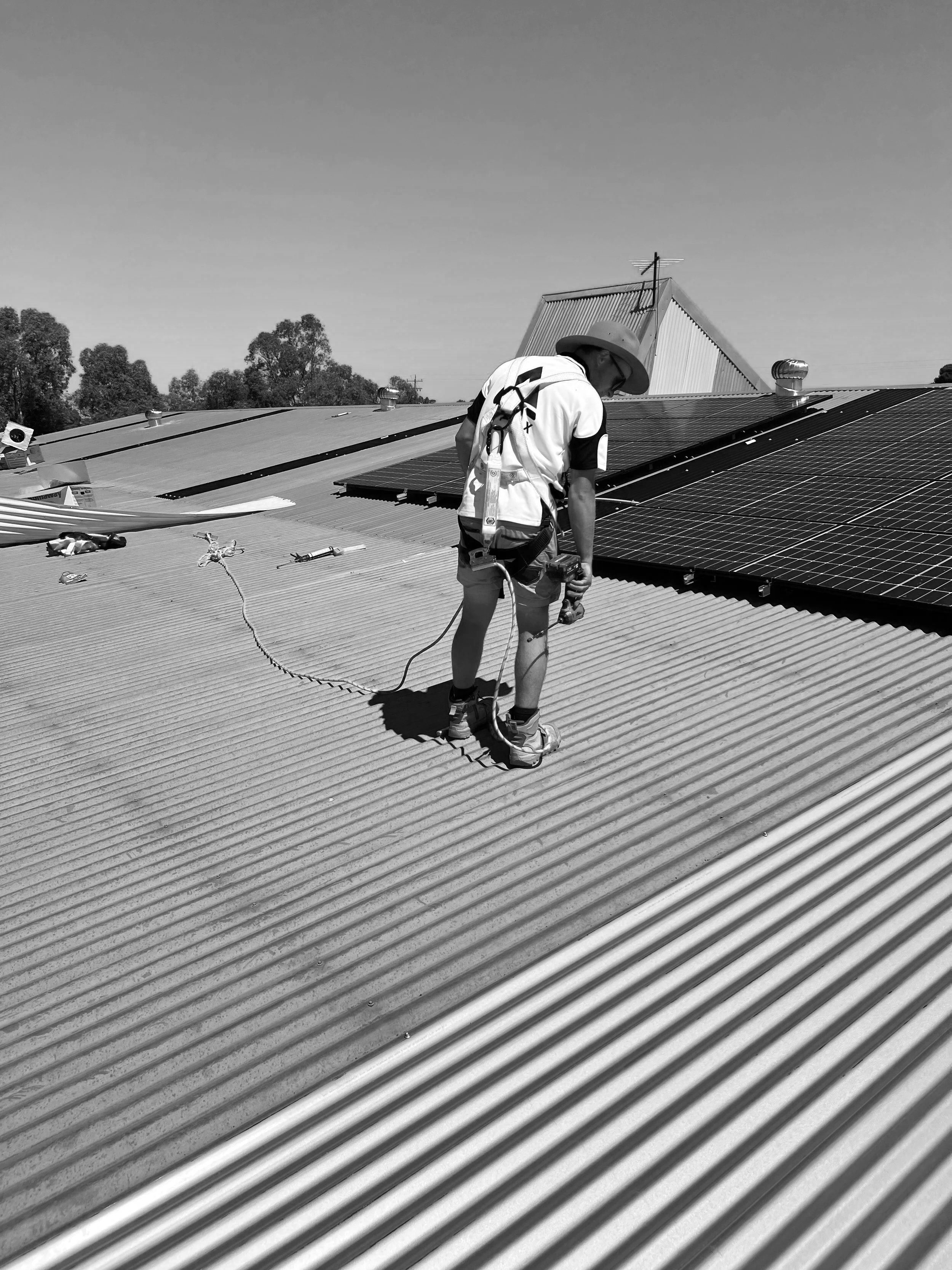 Worker standing on corrugated roof wearing a harness and holding a drill inspecting the roof.