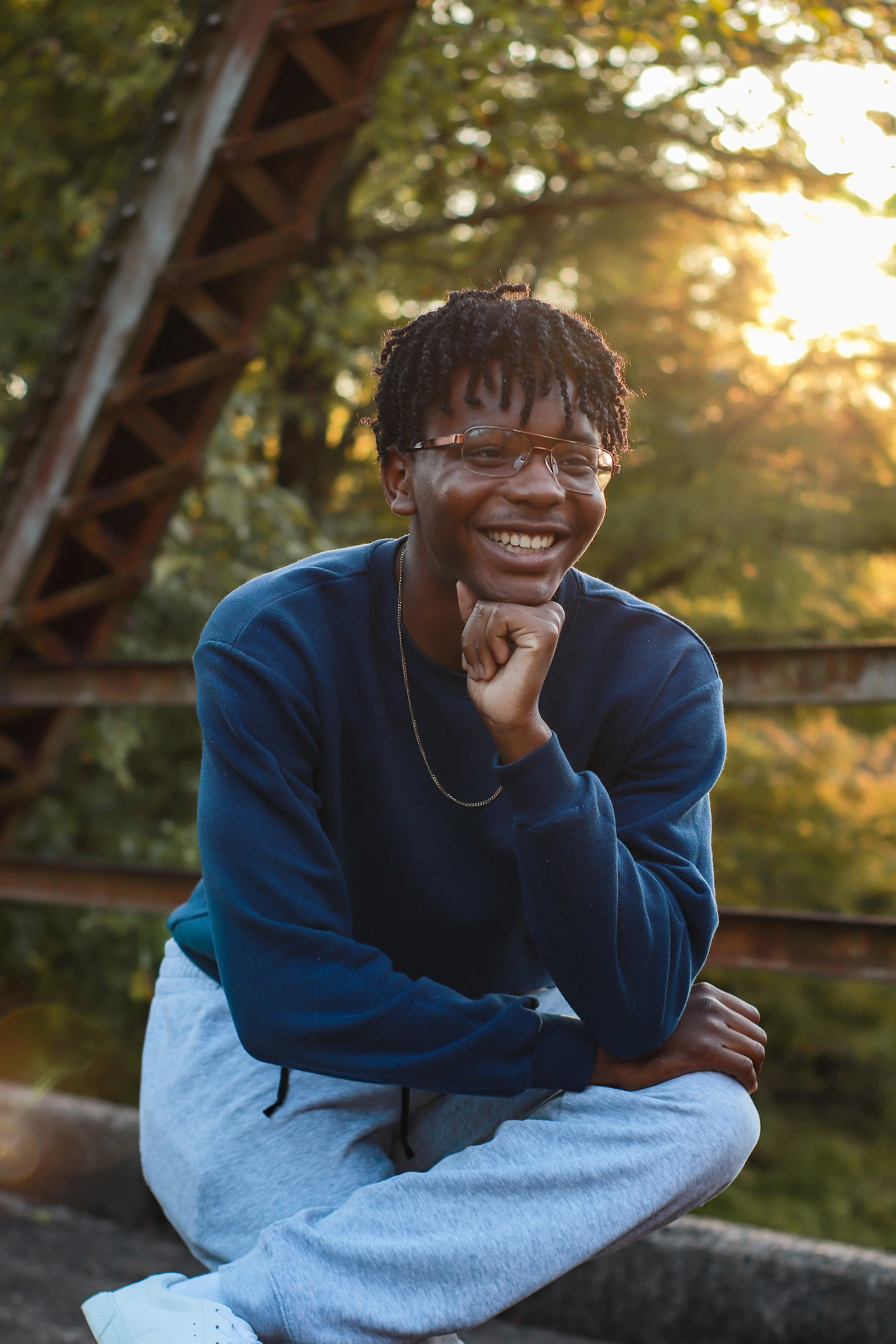 A young man with glasses and dreadlocks smiling outside during sunset, sitting on a rock with a wooden bridge and trees in the background.