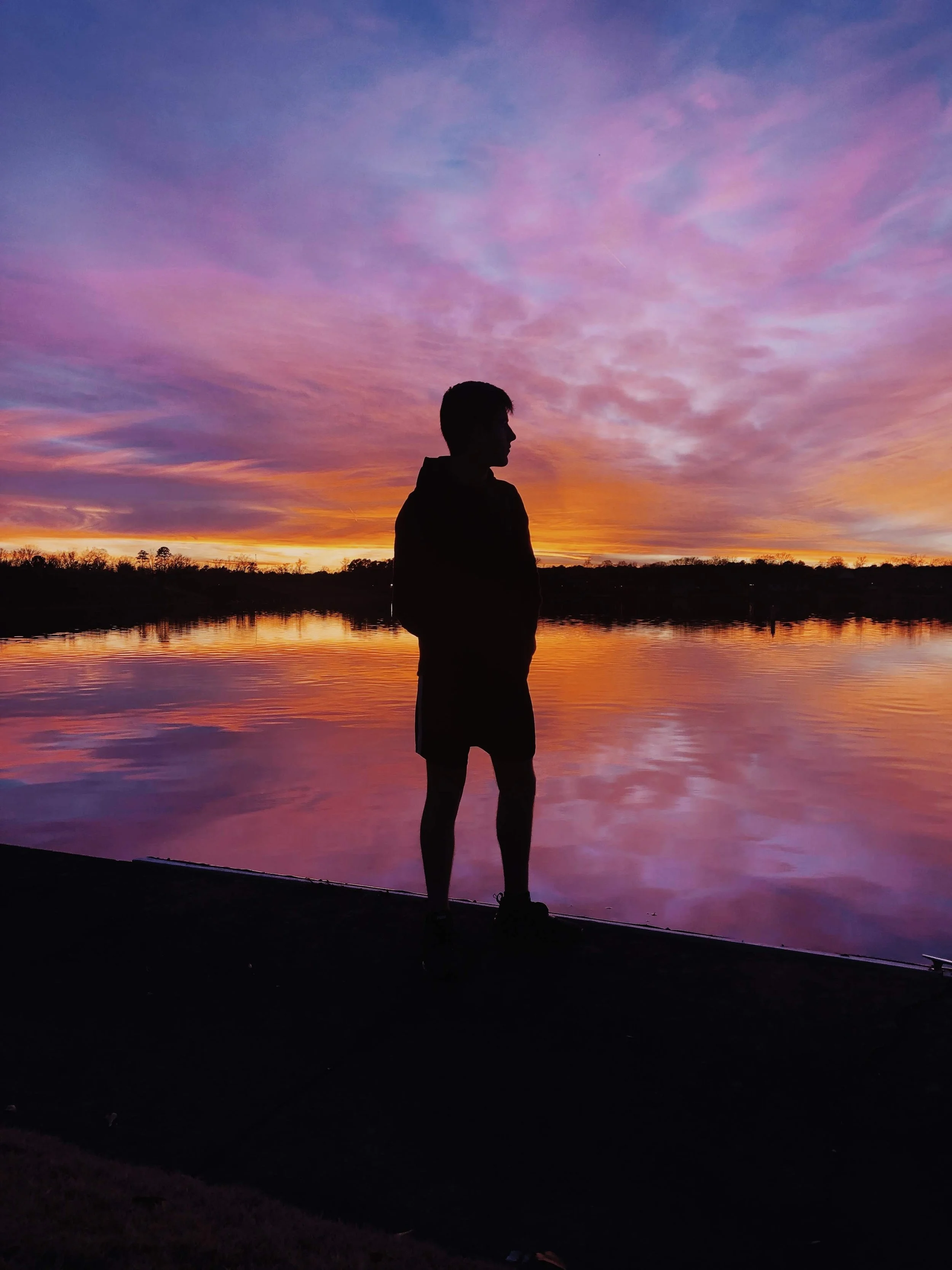 Silhouette of a person standing by a lake at sunset with colorful sky and reflected water.