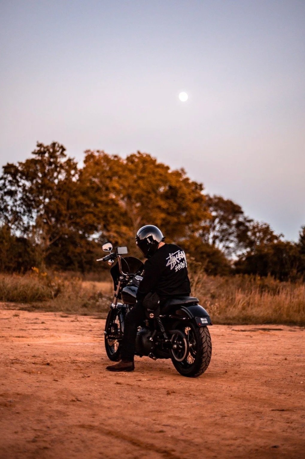 A person wearing a black helmet and black jacket with white text, sitting on a black motorcycle on a dirt path during dusk, with trees and the moon in the background.
