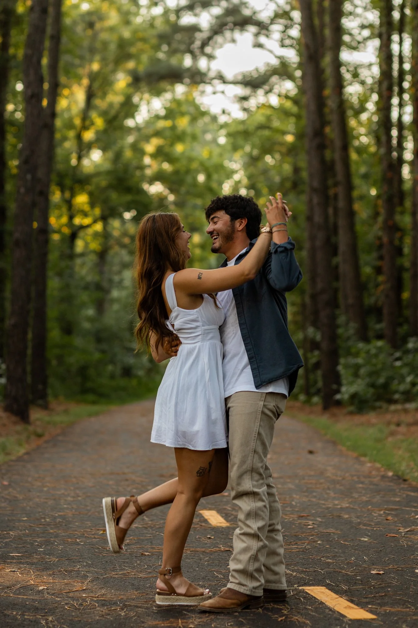 A couple dancing joyfully on a forest path surrounded by tall trees.
