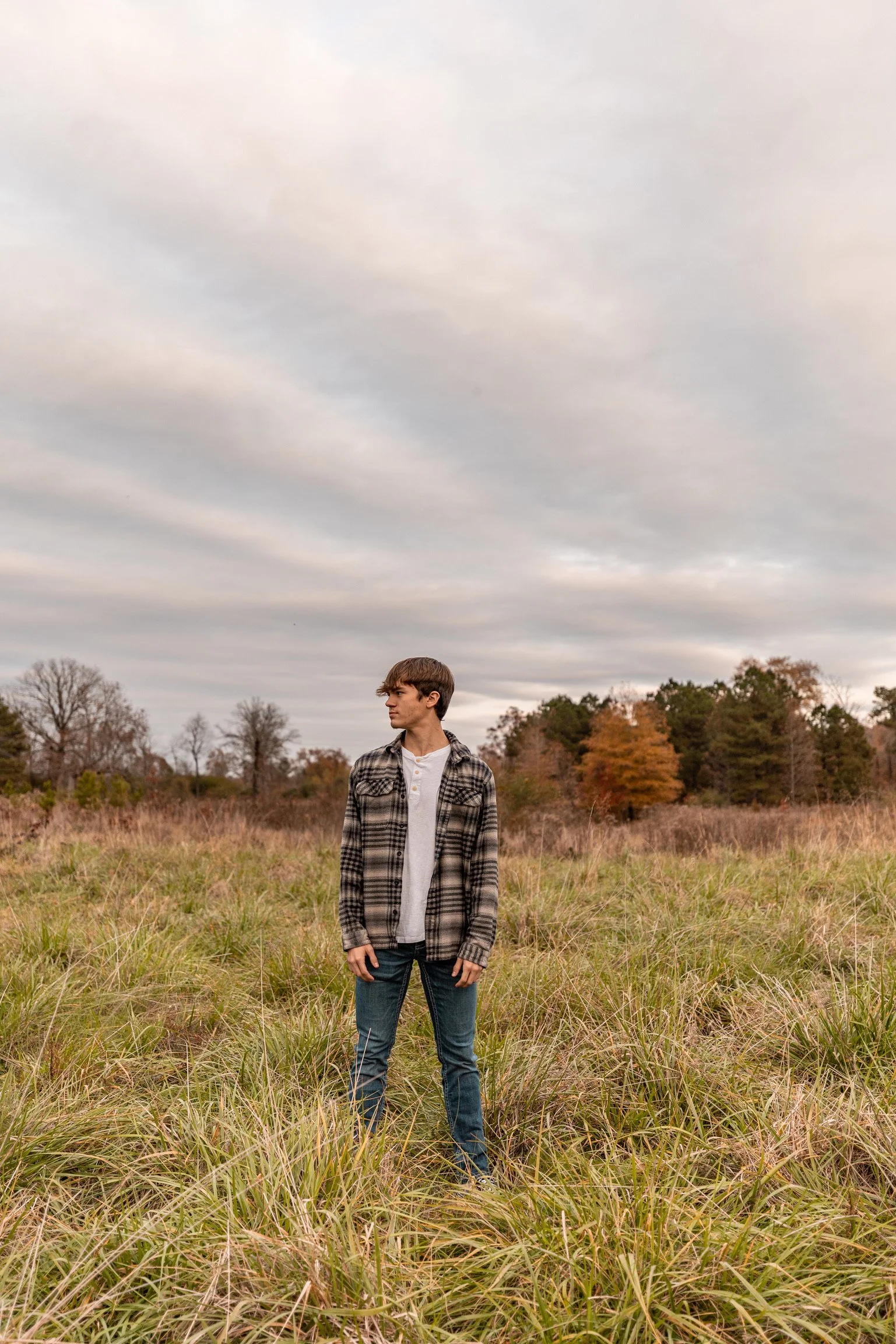 Young man standing in an open grassy field with trees in the background, overcast sky.