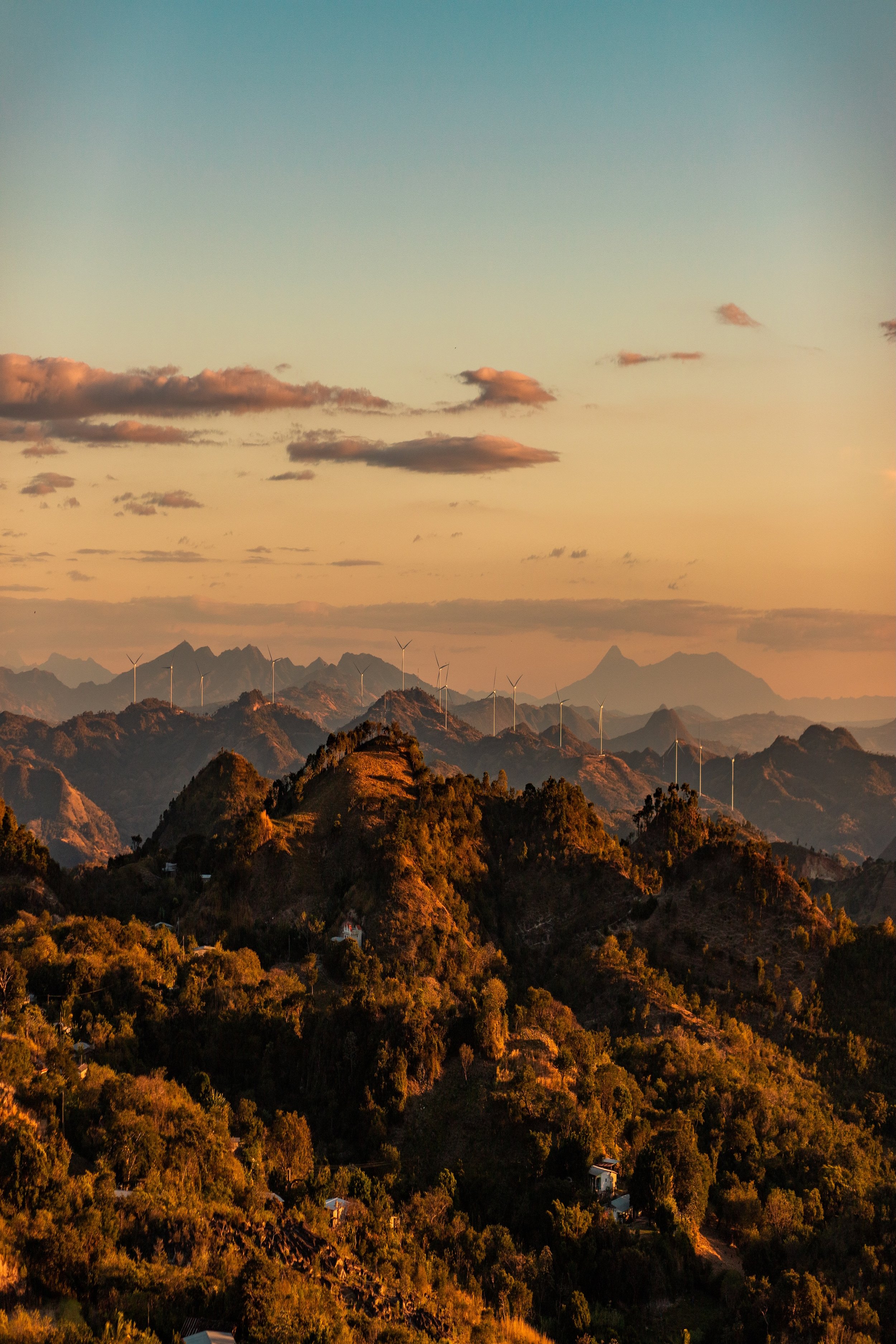 A mountainous landscape at sunset with wind turbines on the ridge and a mostly clear sky with a few clouds.