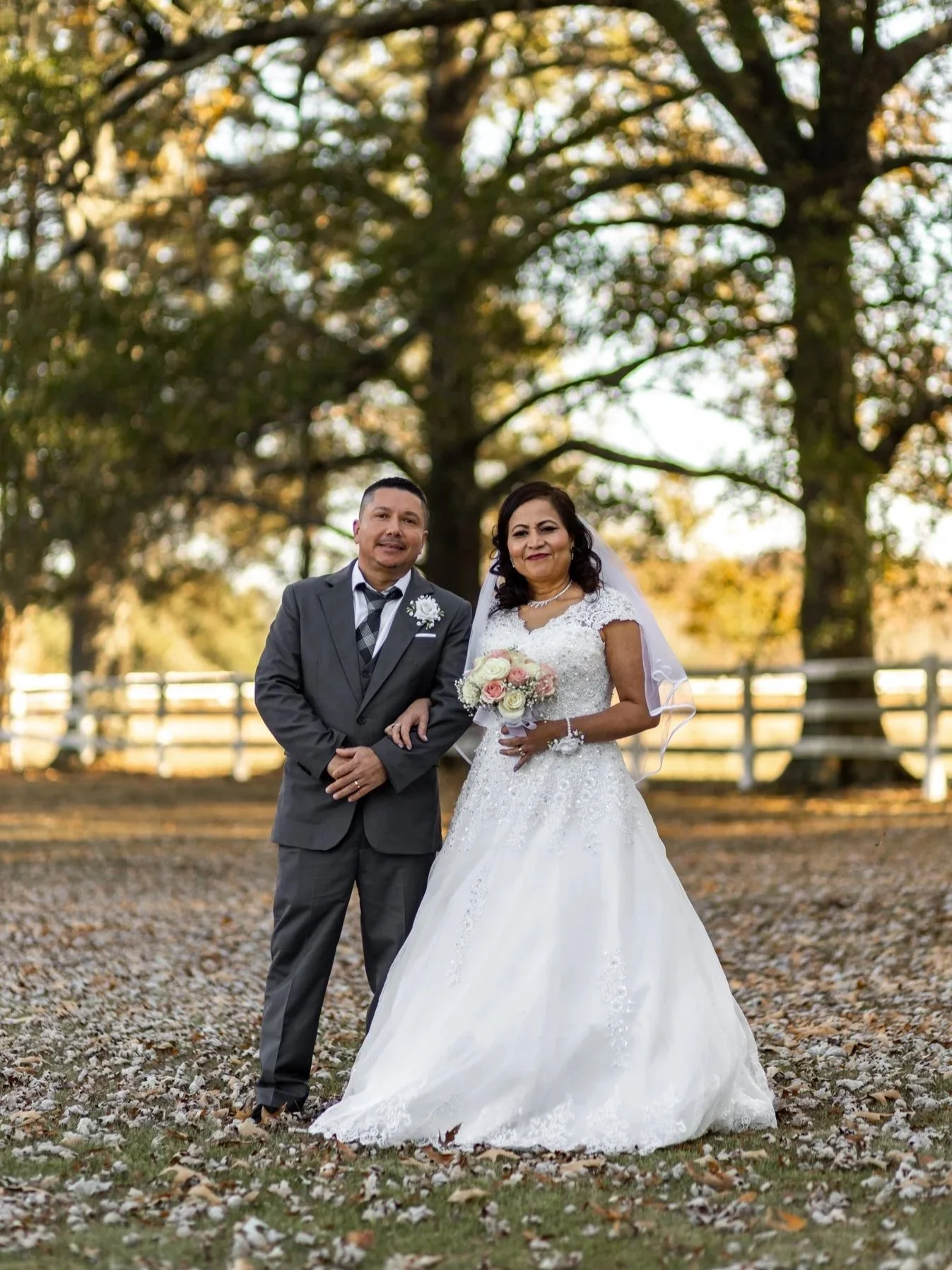 A bride and groom stand outdoors under a large tree, with autumn leaves on the ground, during a wedding celebration in the late afternoon.