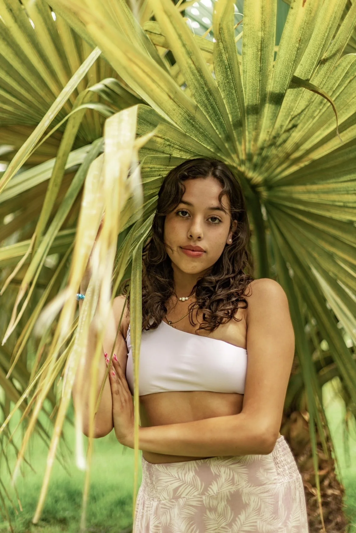 Young woman with curly brown hair and light brown skin standing among large green palm leaves, wearing a white strapless top and patterned skirt, with a neutral expression.