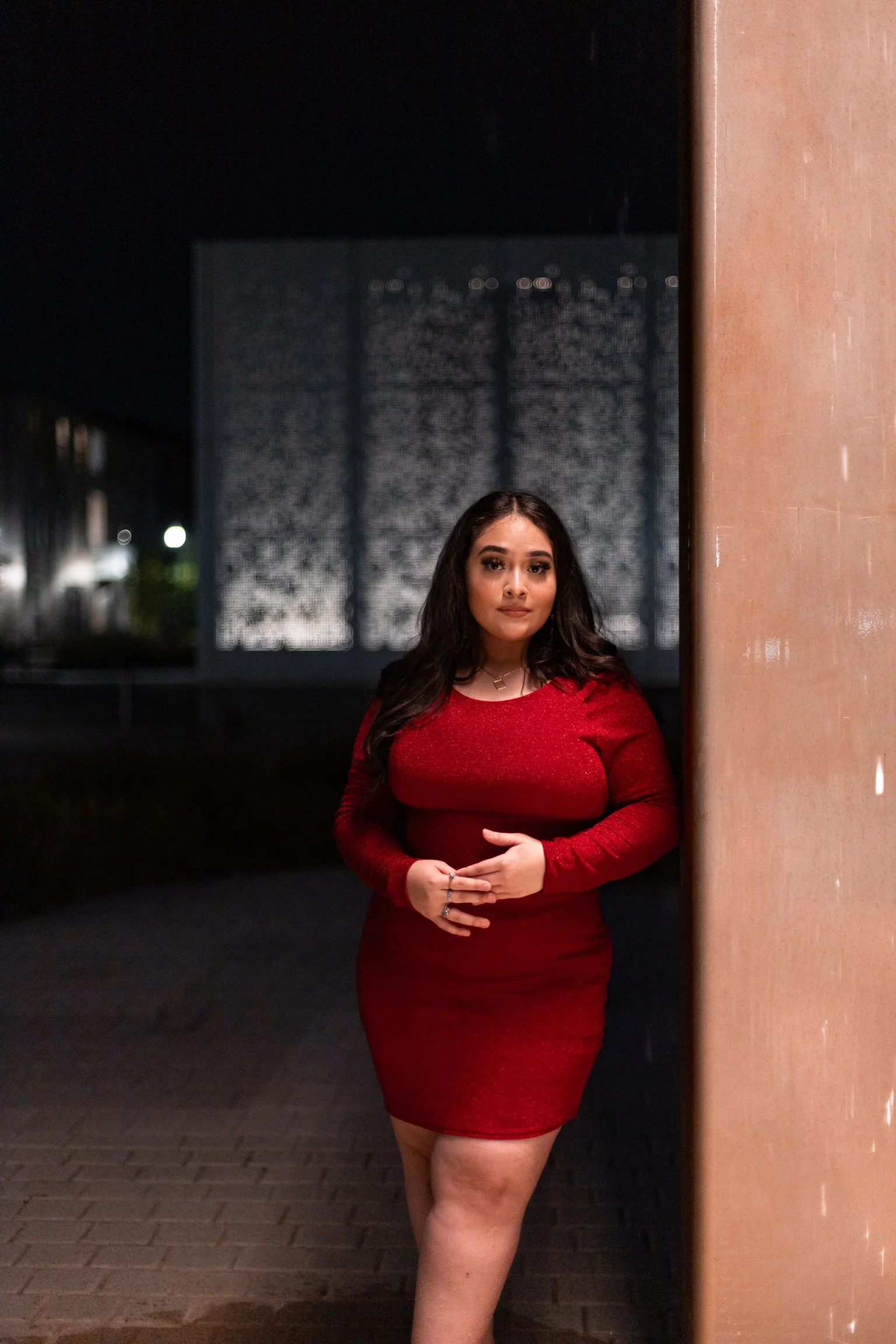 A woman wearing a red dress standing outdoors at night against a background featuring a textured wall and subtle city lights.