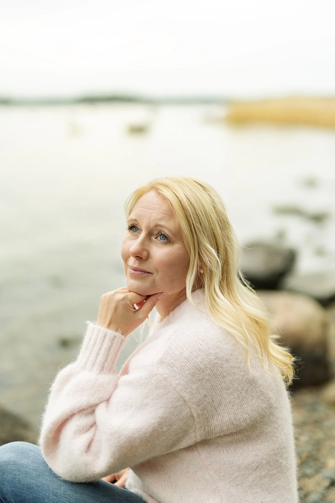 Nainen istuu järven rannalla, katsoo mietiskelevästi taivaalle, taustalla veneitä ja vettä.