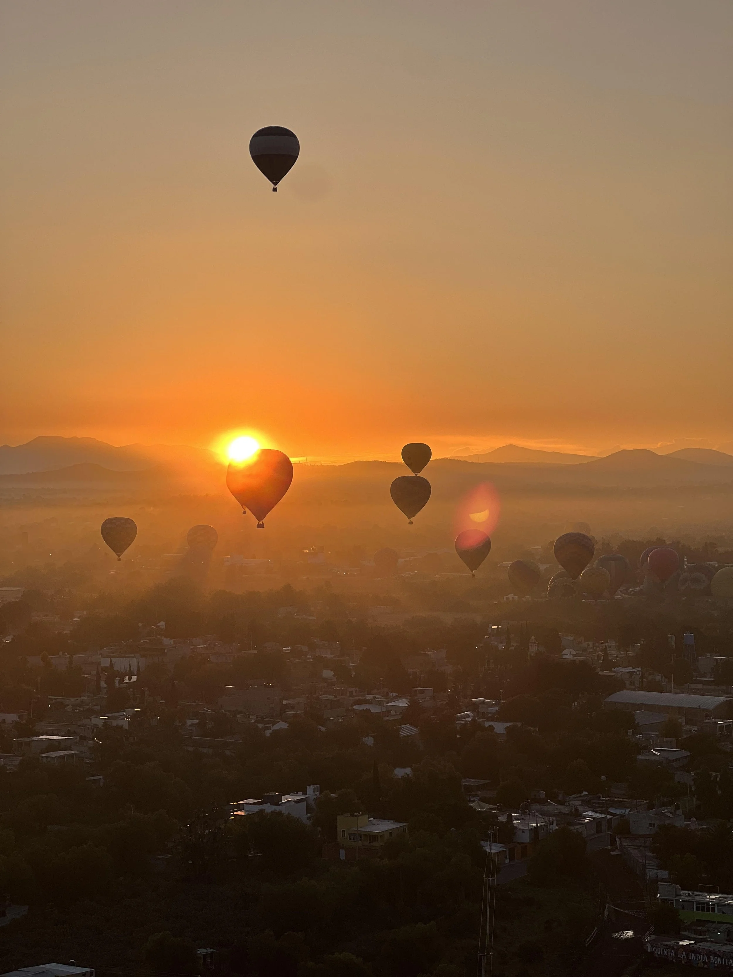 Hot air balloons floating in the sky during a sunset over a town with mountains in the distance.