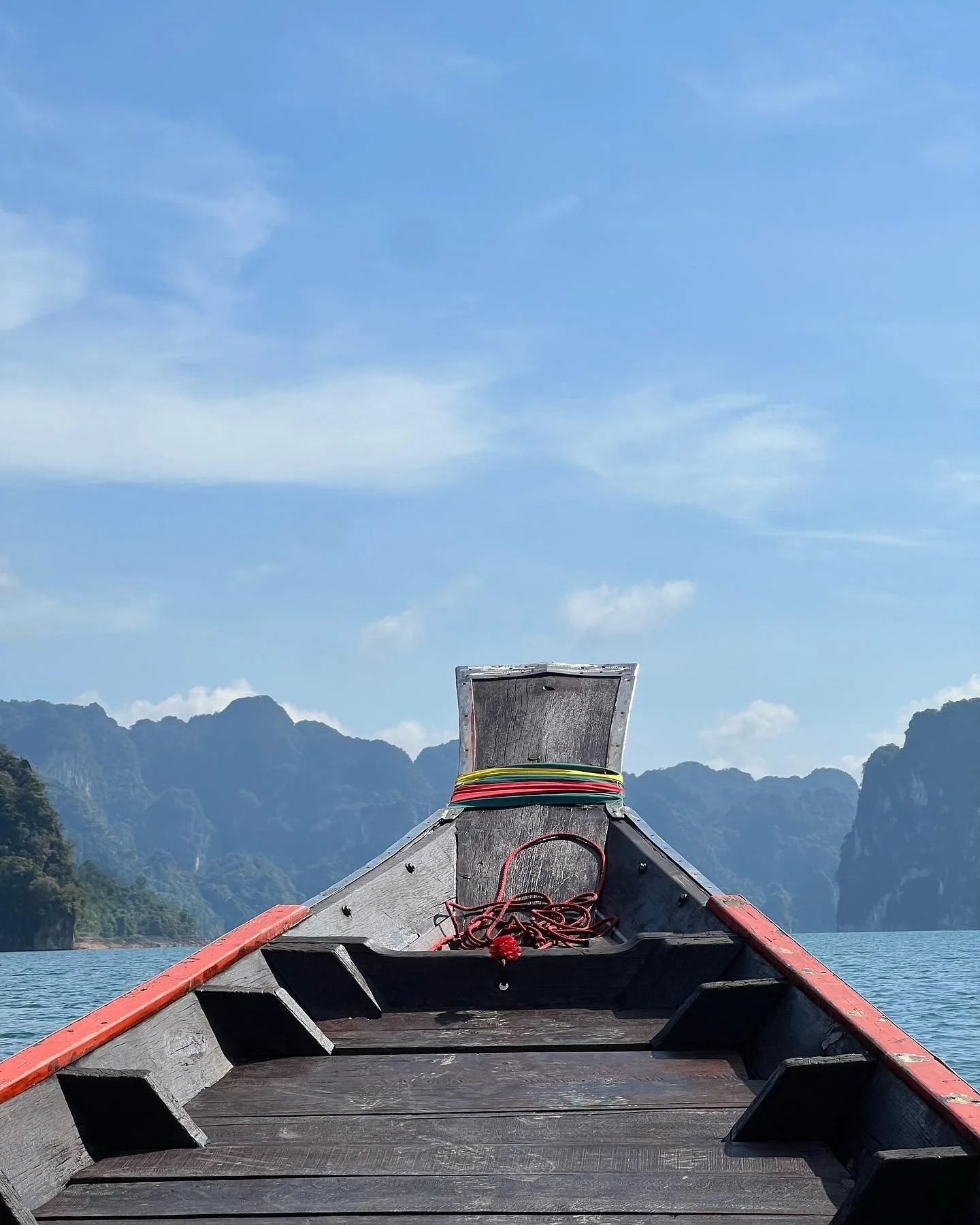 View from the front of a boat with a wooden bow and red trim, floating on a body of water with mountainous landscape and blue sky in the background.