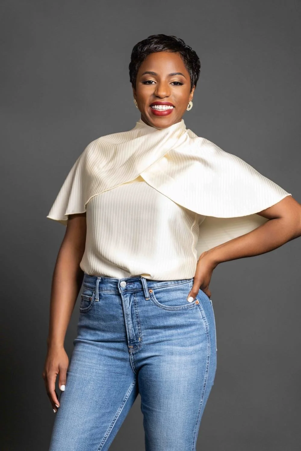 Portrait of a smiling Black woman with short hair, wearing a cream-colored satin blouse and blue jeans against a gray background.