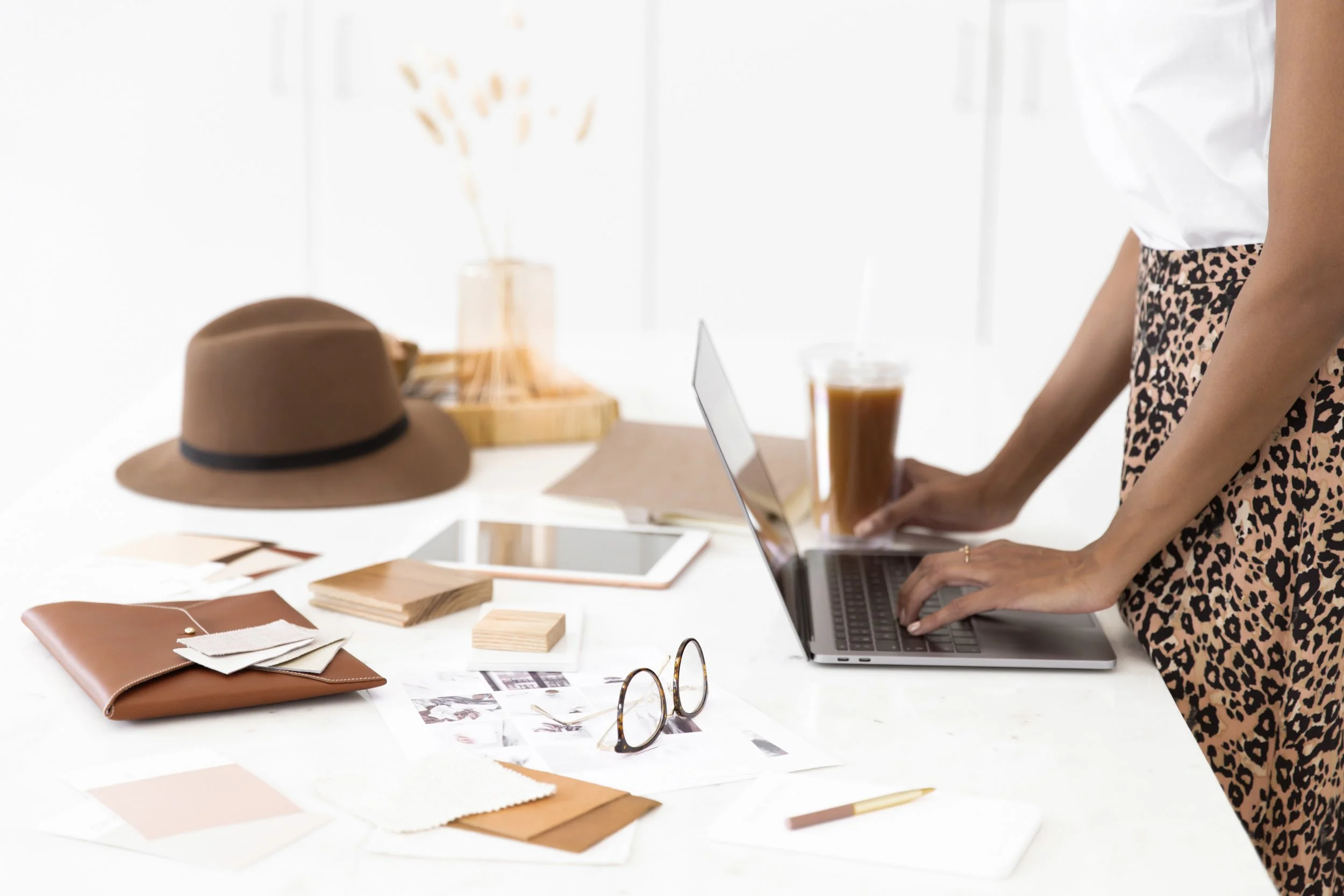 Womanworking at a white desk with fashion design materials, a laptop, a tablet, a drink, and accessories.