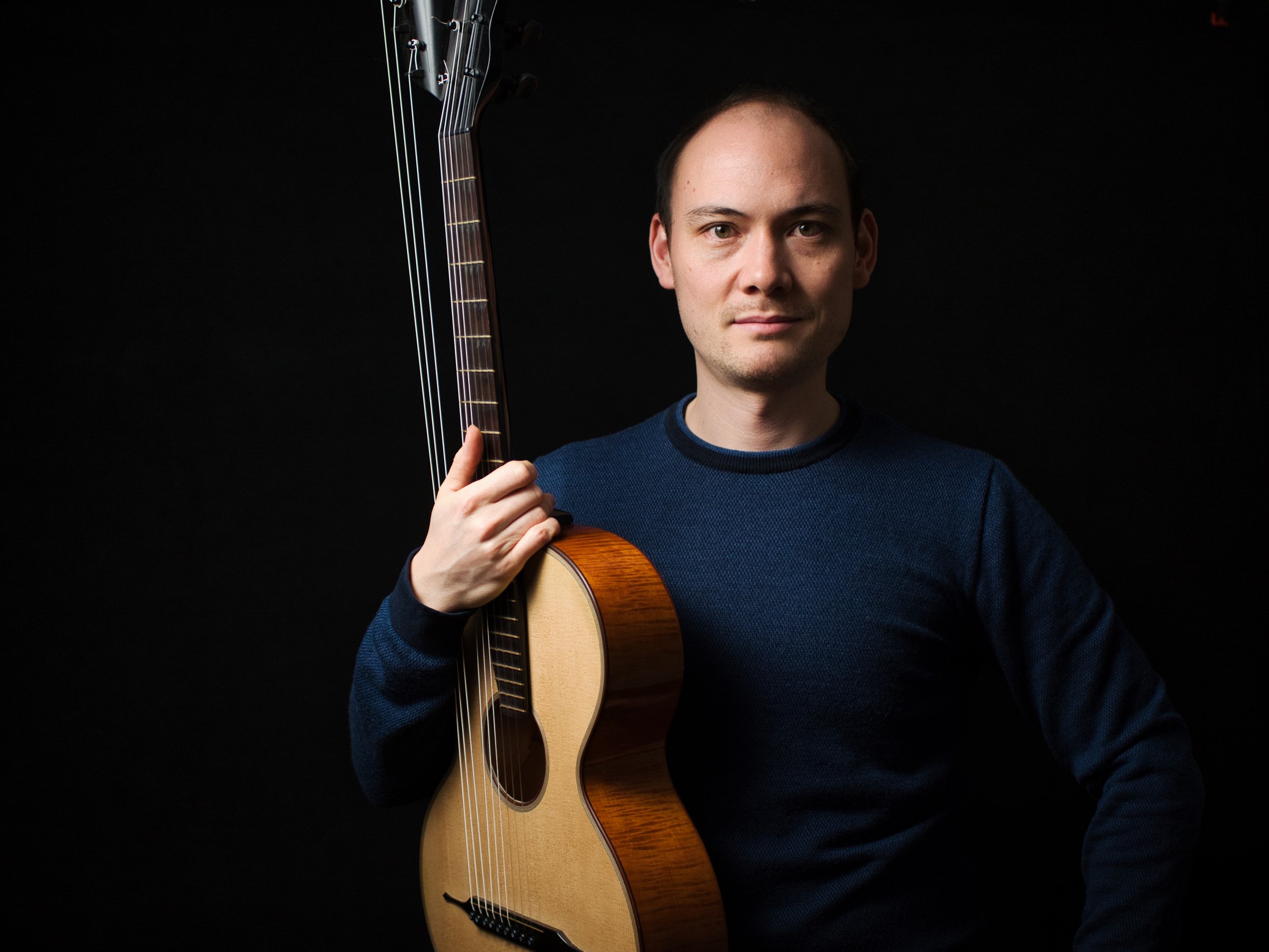 Mann mit dunklem Pullover hält eine Akustikgitarre vor dunklem Hintergrund.