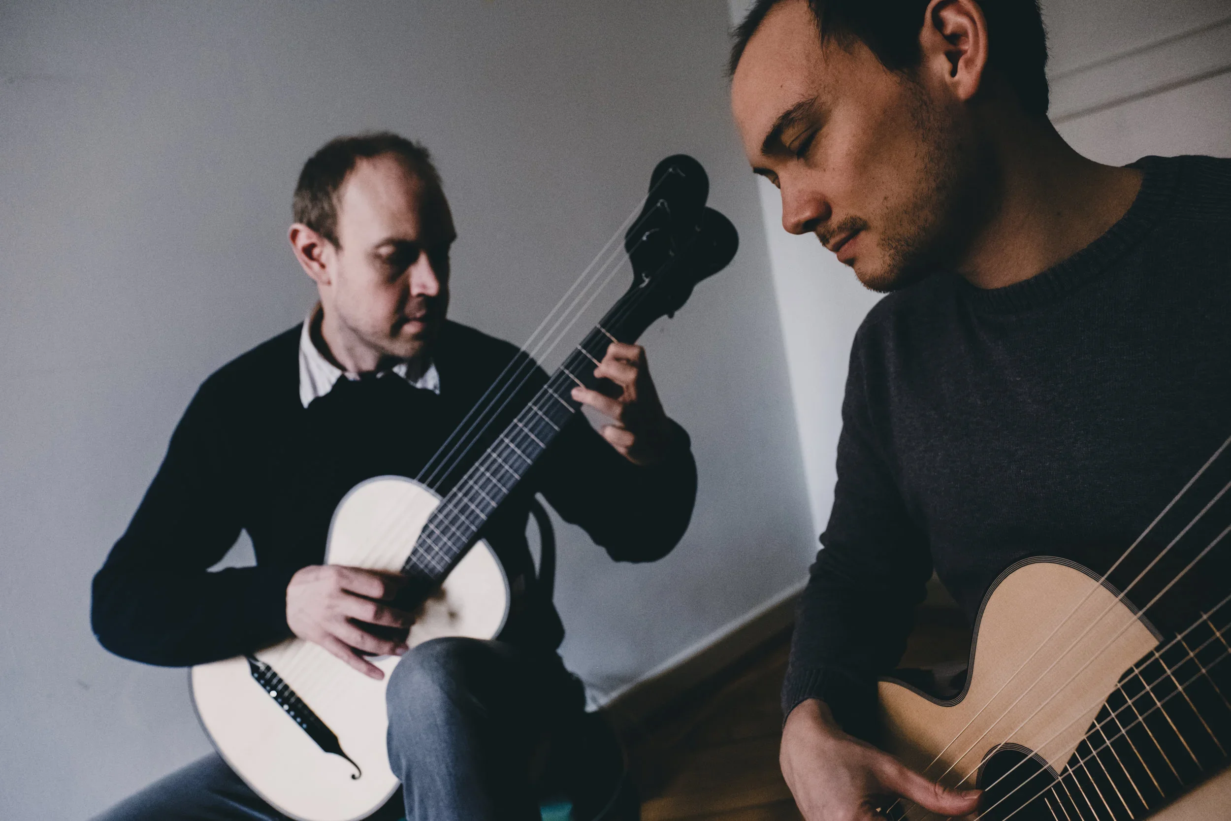 Zwei Männer spielen Gitarre in einem Raum, einer sitzt mit einem E-Gitarrenkopf nach vorne und der andere mit einem Akustik-Gitarre, beide sind konzentriert auf ihr Instrument.