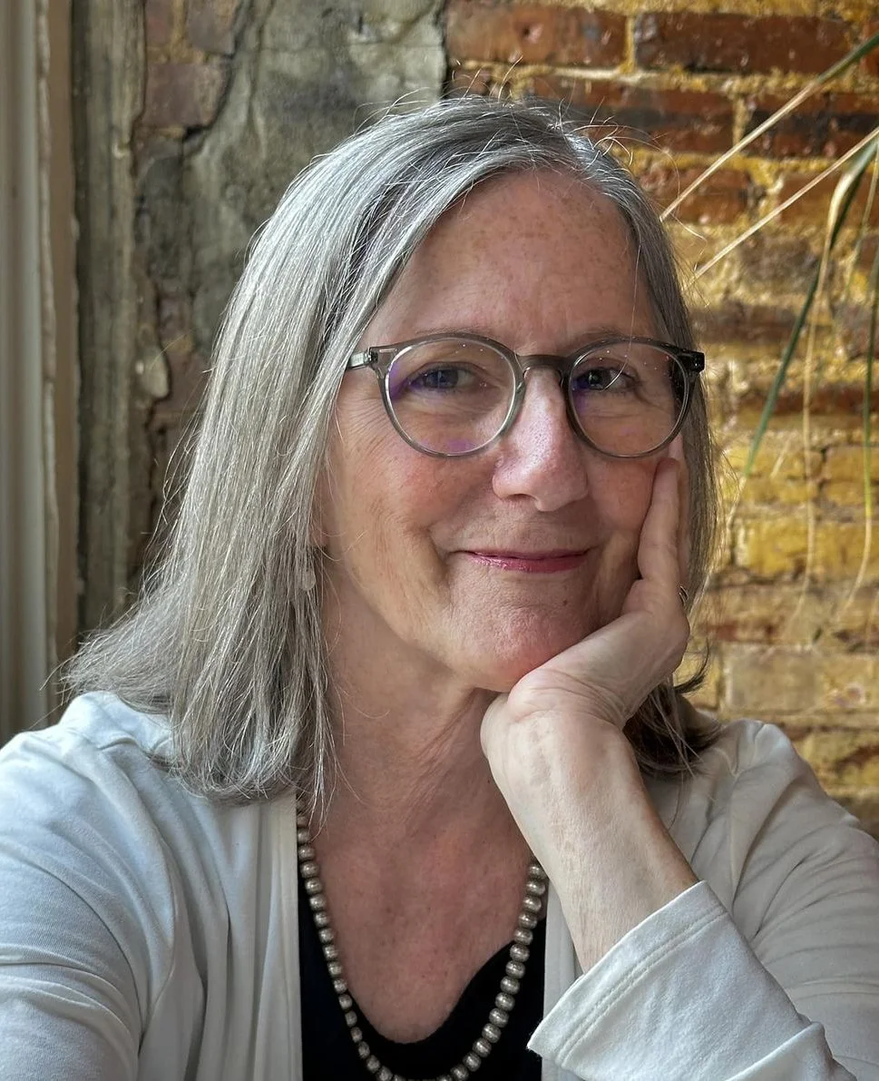 A woman with gray hair, wearing glasses, a black top, a pearl necklace, and a white cardigan, resting her chin on her hand, smiling softly in front of an exposed brick wall.