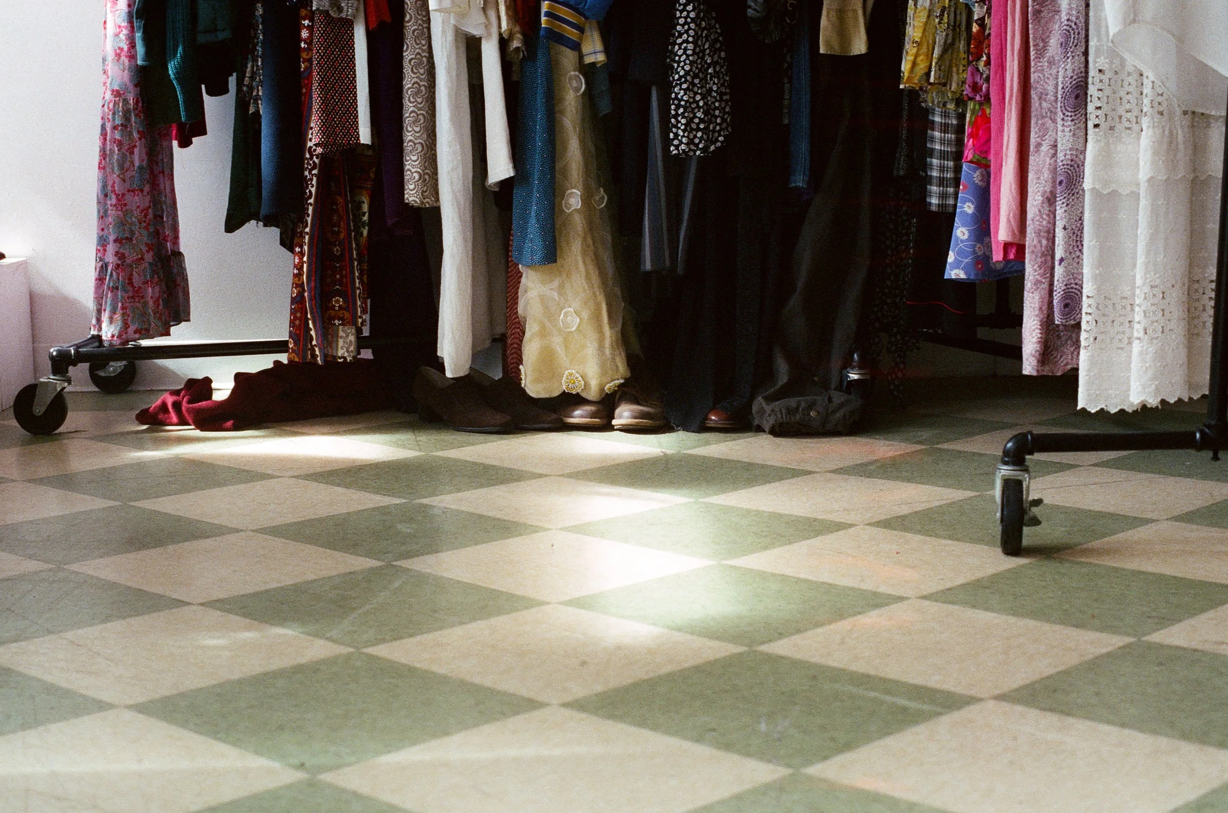 Clothing rack with assorted dresses and garments, some shoes on the floor, in a room with checkered tile flooring.
