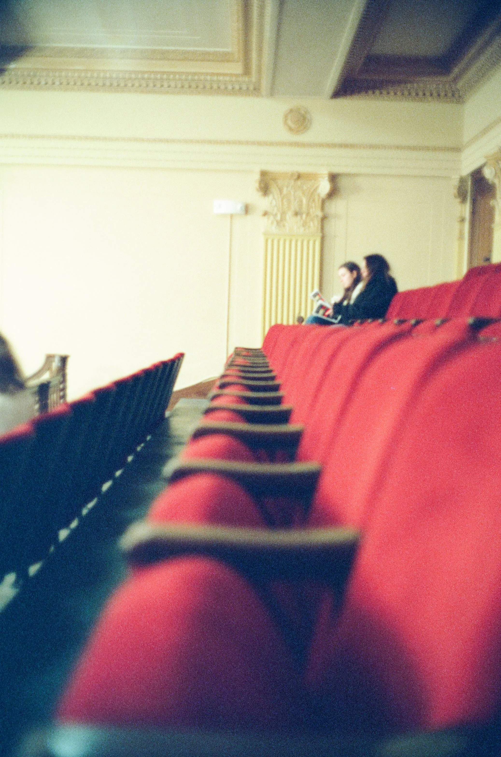 Empty theater seats with a few people sitting in the back row, reading a book or looking at a device, ornate ceiling details visible.