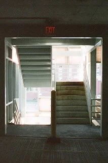 Indoor stairway leading upward, illuminated by natural light from large windows, with an exit sign above the door.