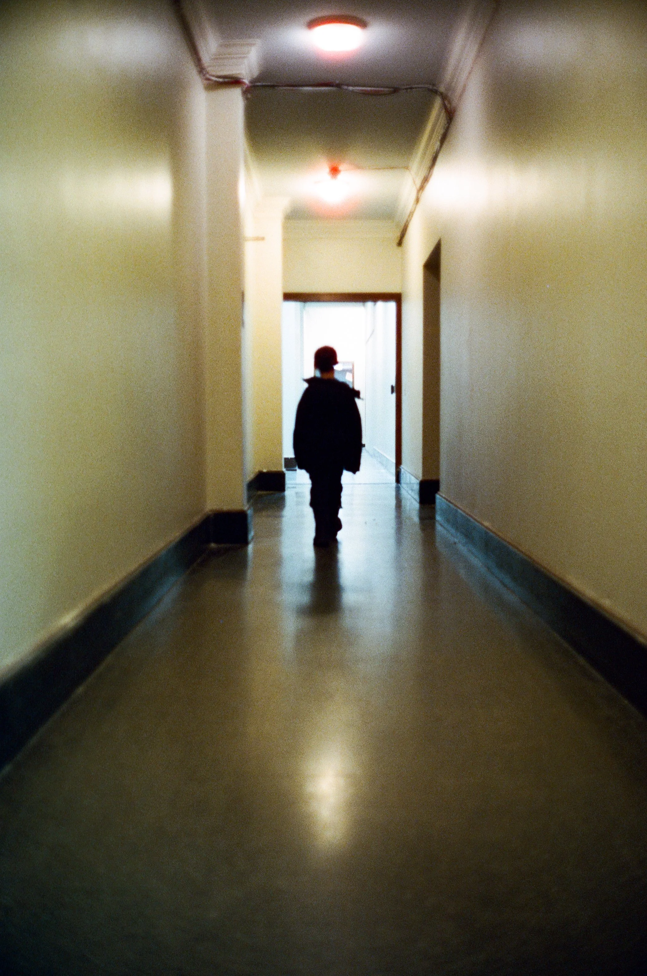 A person walking down a dimly lit hospital corridor with beige walls and dark flooring, towards a brighter area at the end.