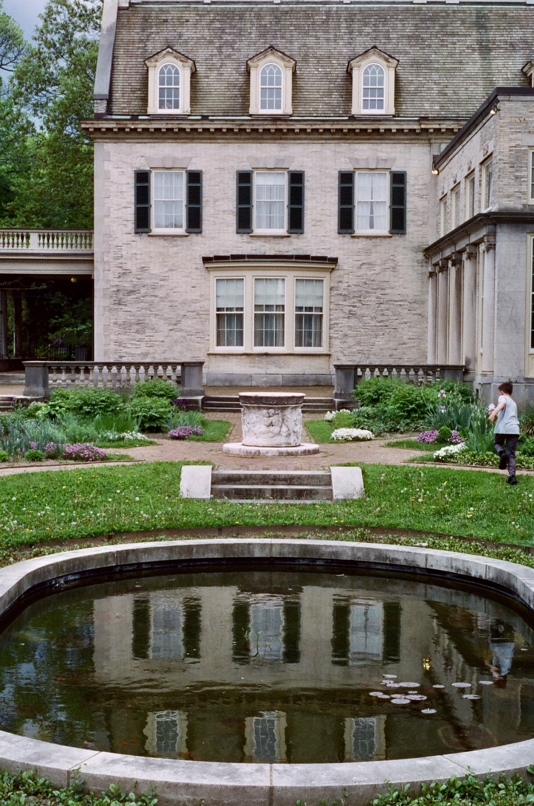 Front yard of a large brick house with a fountain, garden, and a young boy walking along a flower garden path.