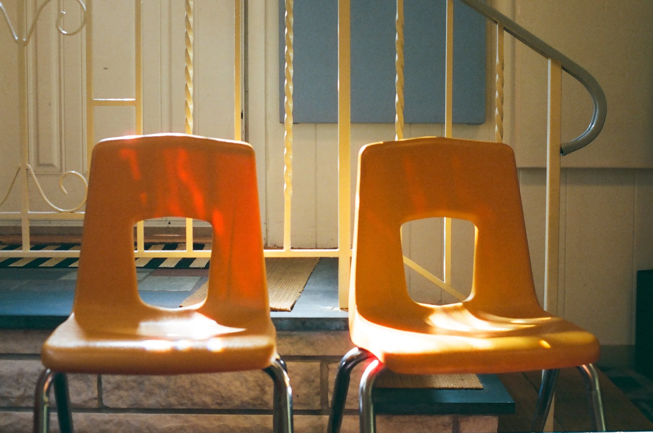 Two yellow plastic chairs positioned in front of a staircase with decorative railings, with sunlight casting shadows on them.