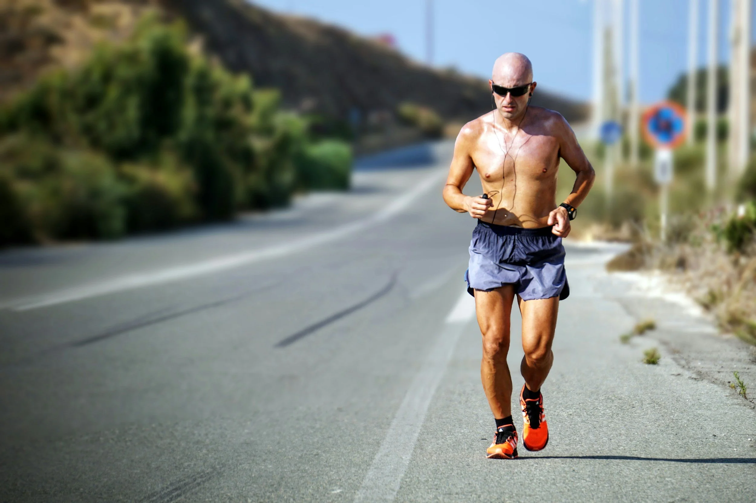 A shirtless man with sunglasses running on the side of a rural road wearing shorts and running shoes.