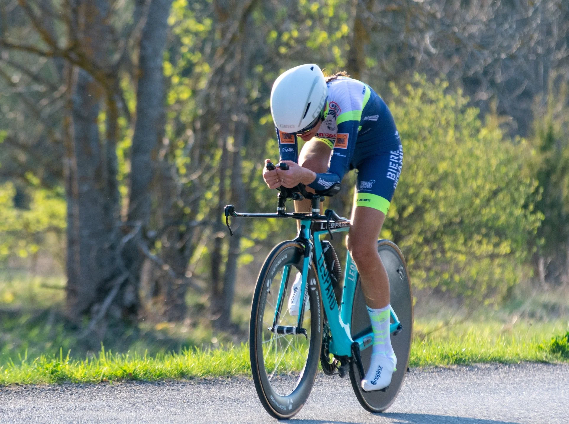 A cyclist wearing a white helmet and a blue and yellow racing suit riding a teal bike on a paved path surrounded by trees and greenery.