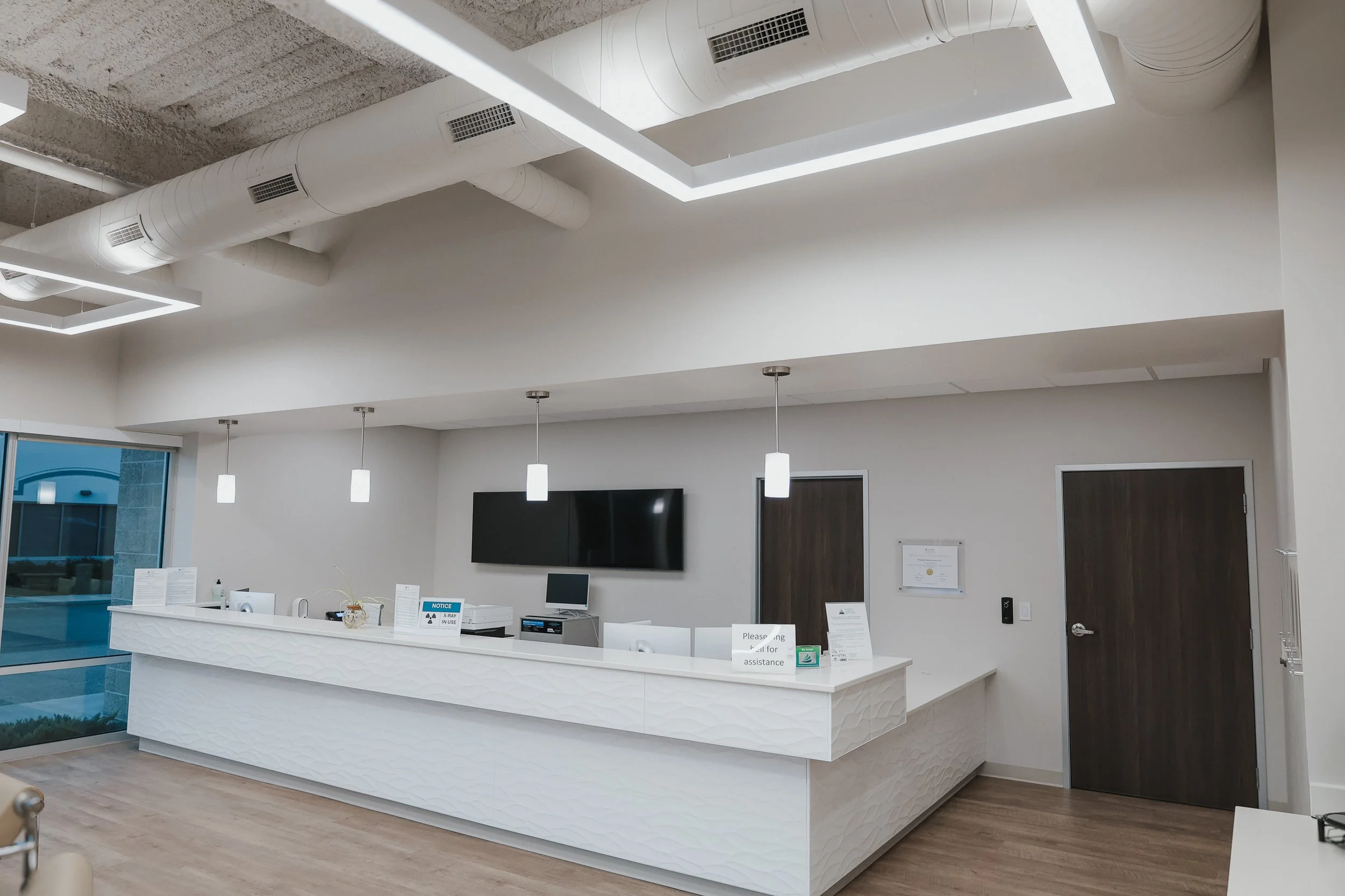 Modern reception area with white textured counter, pendant lights, flat-screen TV, computer, and signs, inside a building with wood flooring, a large window, and ventilation ducts on the ceiling.