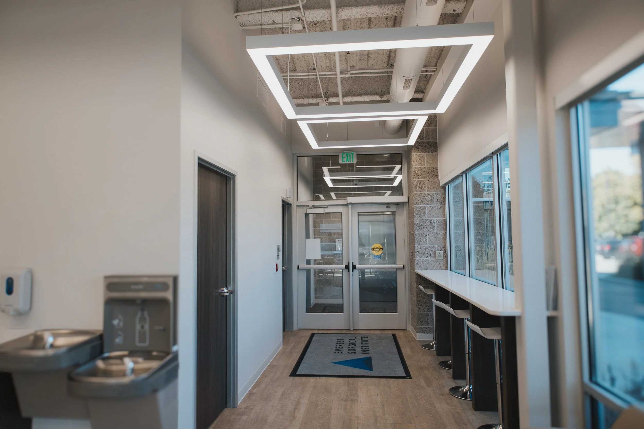 Interior view of a medical office or clinic with water fountain, chairs, and large windows, with a geometric ceiling light design and an exit sign.