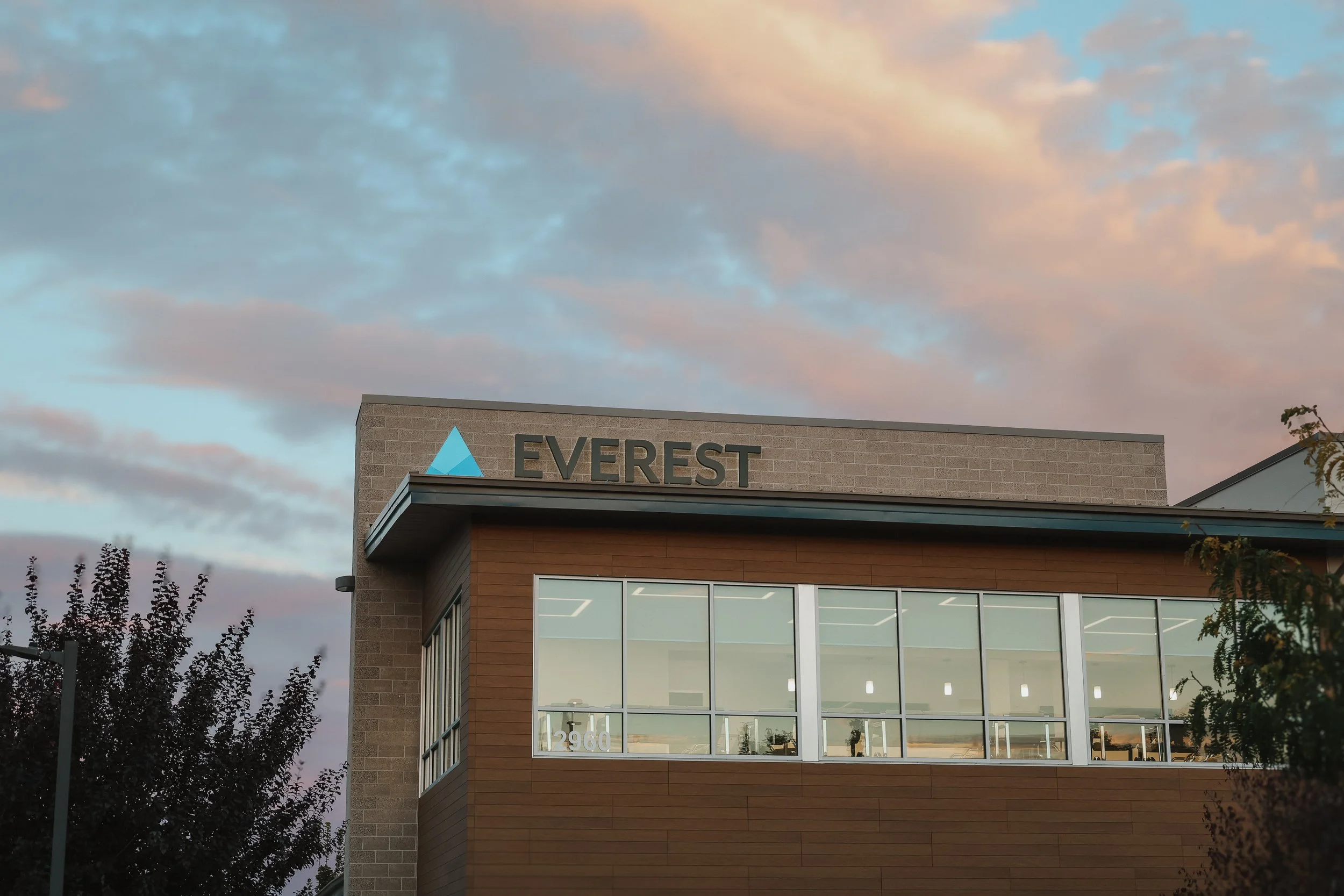 Modern two-story building with a sign that reads 'Everest' and a blue mountain logo on the upper corner, large windows on the second floor, trees, and a colorful sky with clouds at sunset.