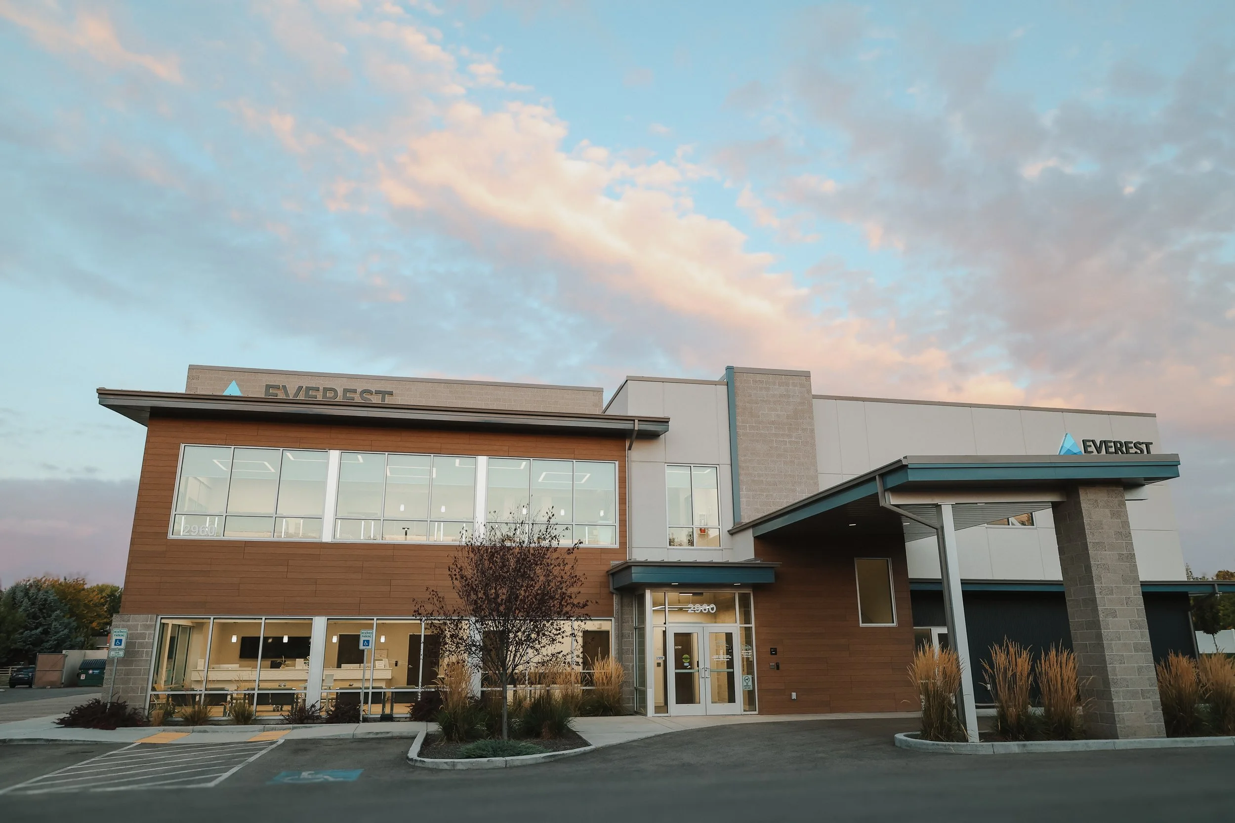 Modern two-story office building with the name Everest, large glass windows, and a parking lot in front with accessible spaces, surrounded by trees and shrubs, under a cloudy sky at sunset.