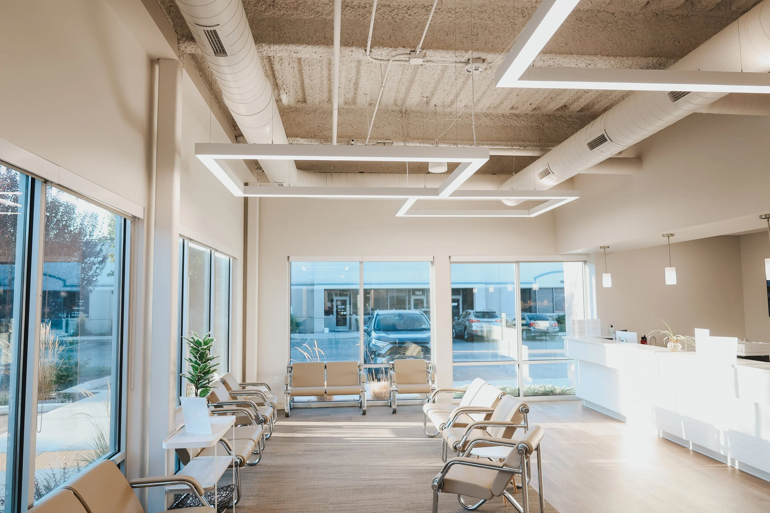 A modern, well-lit waiting room with glass windows, beige chairs, a reception desk, and ceiling lights.