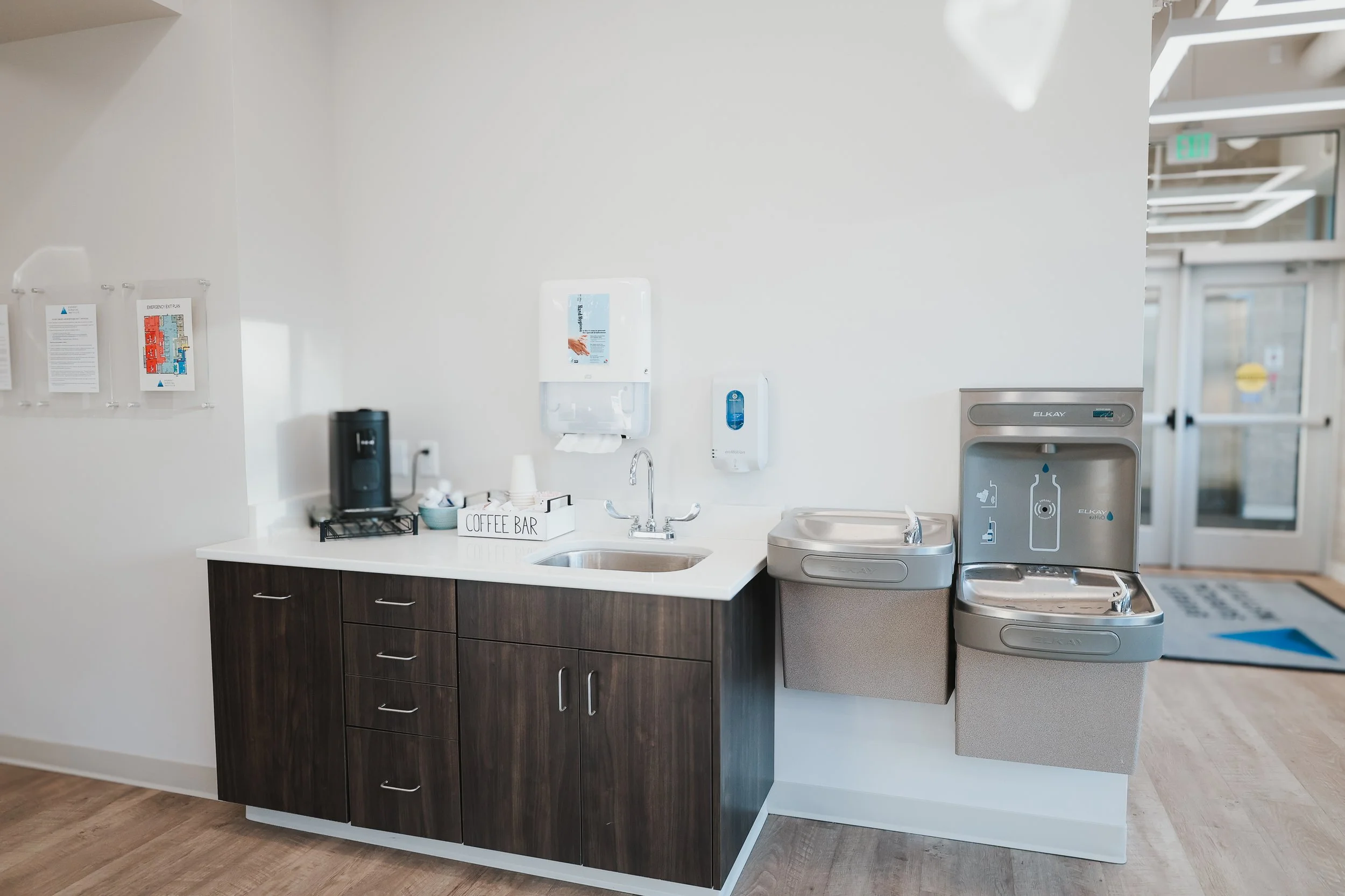 Coffee station with a coffee maker, cups, and a sign labeled 'Coffee Bar' on a countertop with a sink, and a water fountain in a hospital or medical facility.