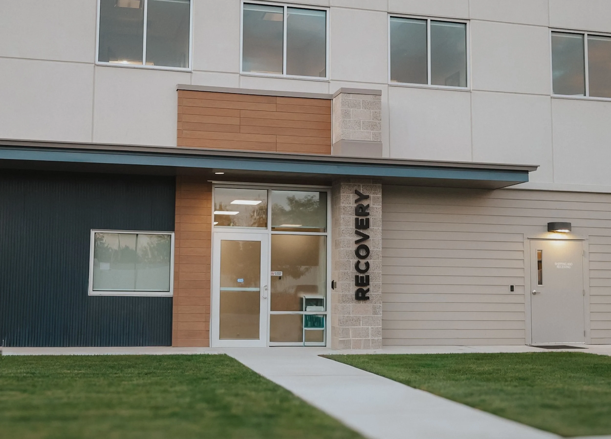 Exterior of a modern building with a glass door labeled 'RECEIVING' and a sign that reads 'RECOUVER' next to the entrance, grass lawn, and a sidewalk leading up to the door.