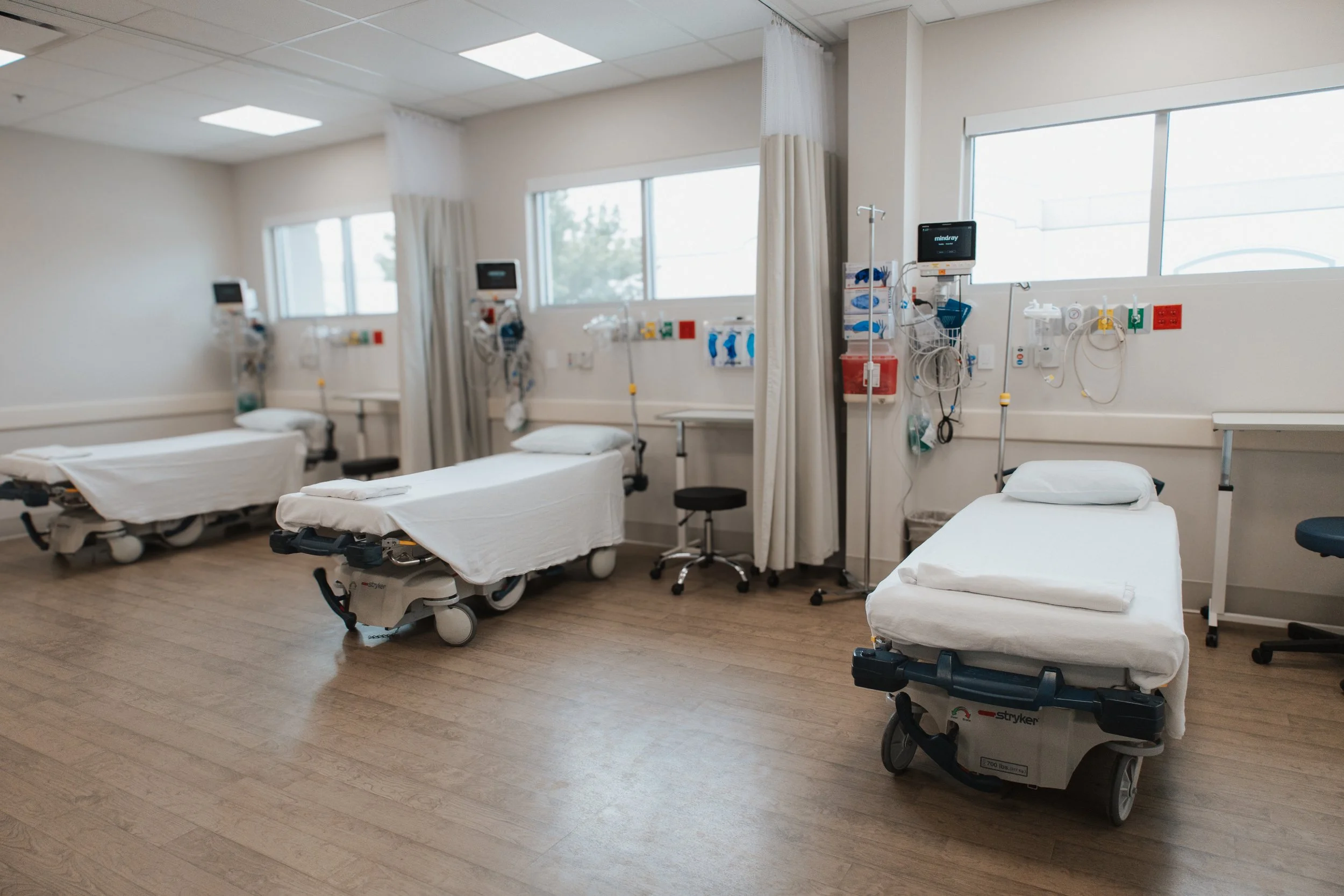 Empty hospital room with three hospital beds, medical monitors, and equipment, windows letting in natural light, beige walls, wooden floor.