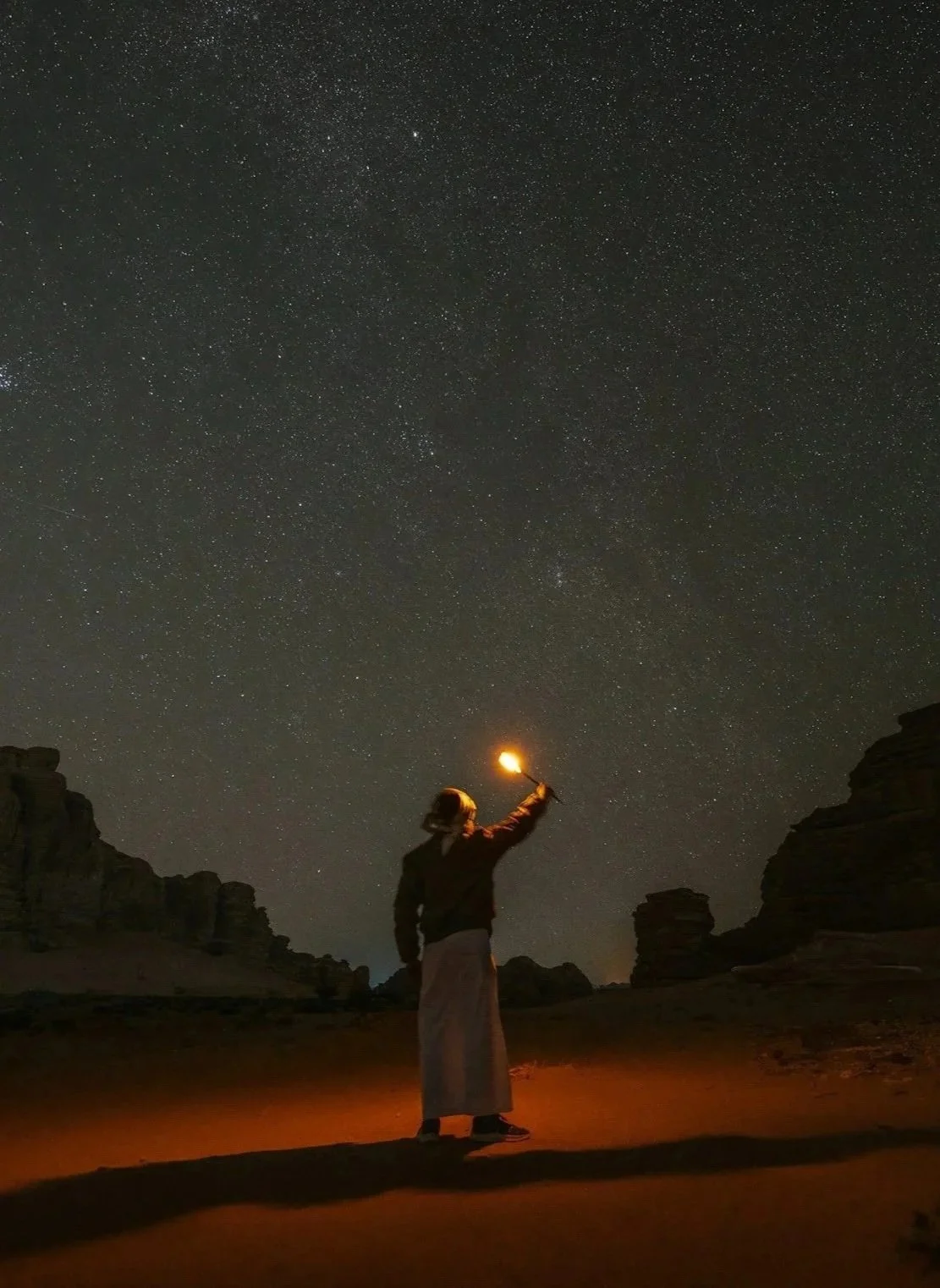 Person standing outdoors at night, holding a small torch pointed towards the starry sky, with rocky formations in the background.