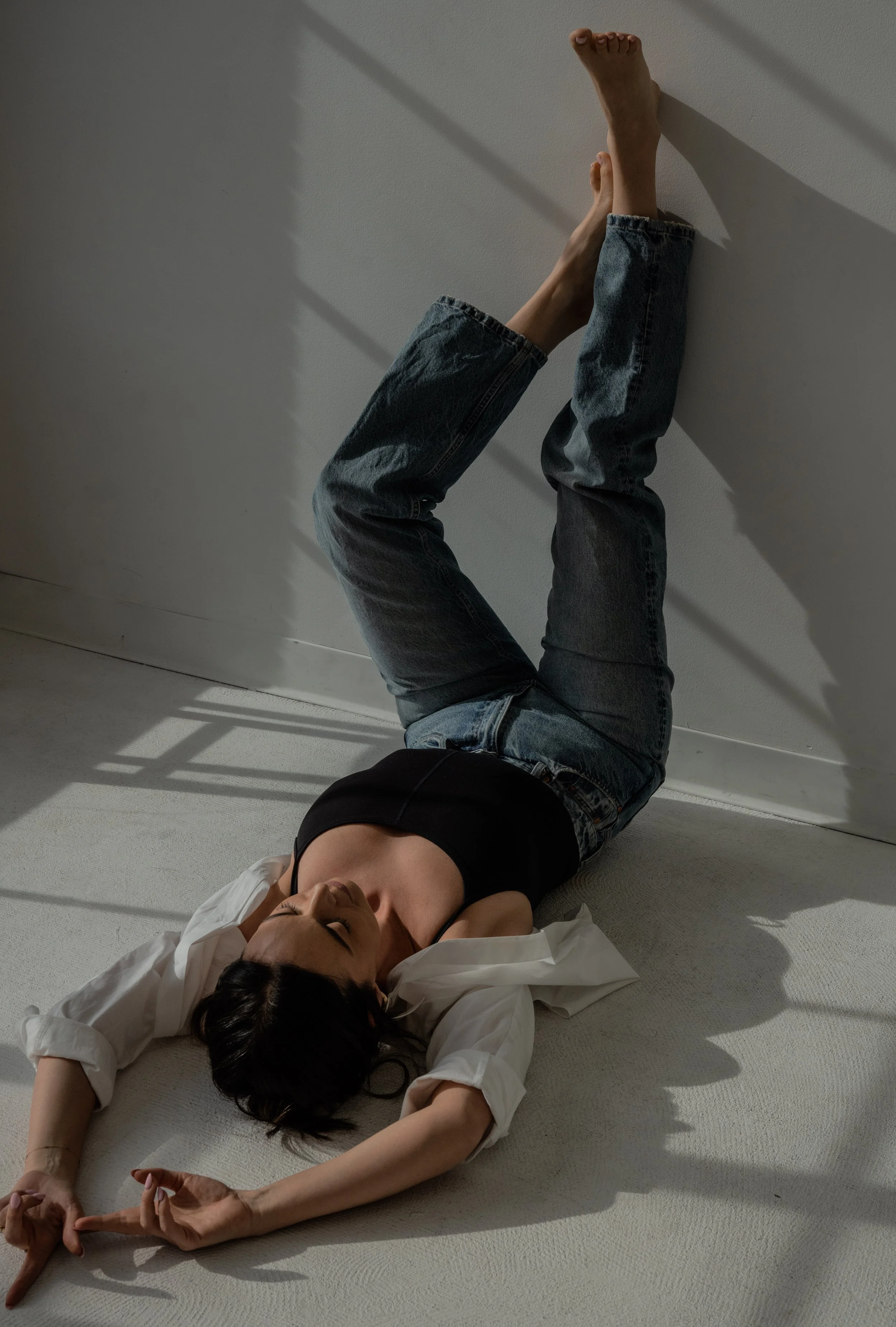 A woman lying on the floor with her eyes closed, wearing a black tank top, white shirt, and jeans, stretching her arms above her head and touching her toes, with sunlight casting shadows on the wall.